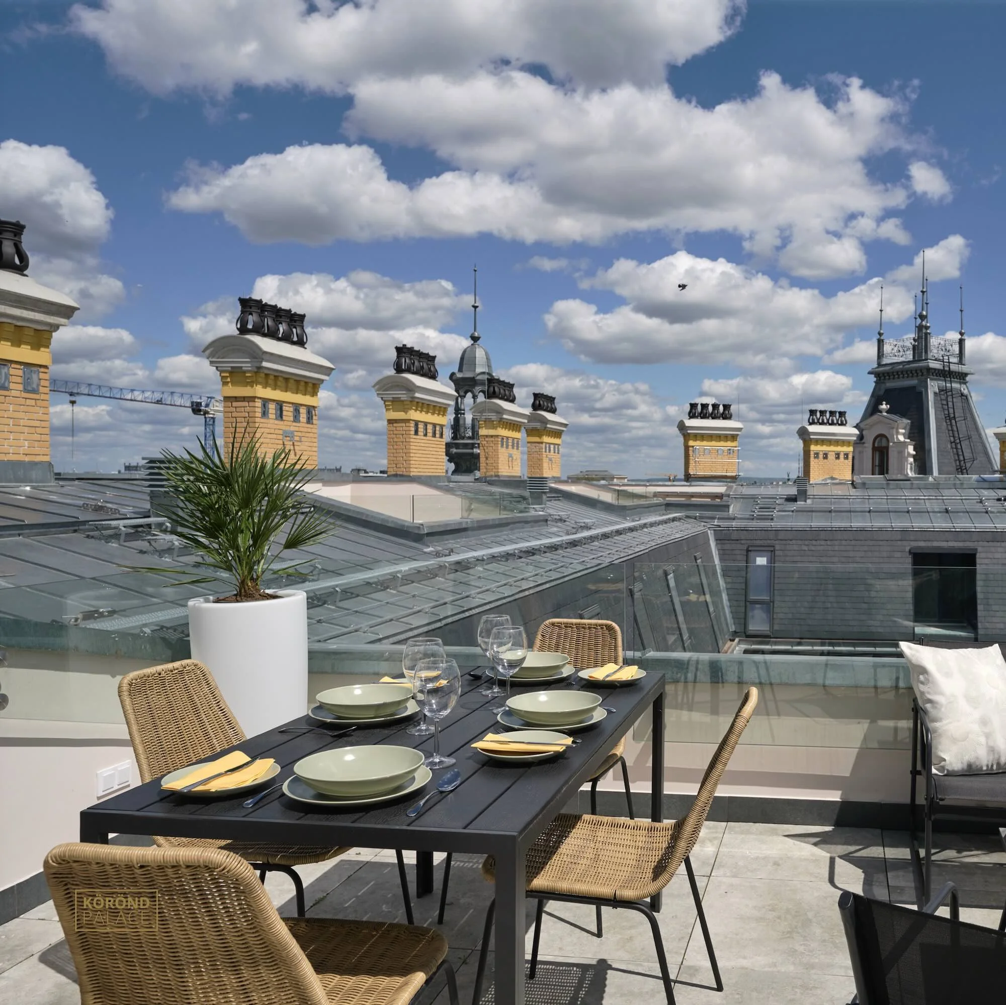 Rooftop dining area with a table set with plates, glasses, and napkins, surrounded by wicker chairs, overlooking city rooftops and a bright, cloudy sky.