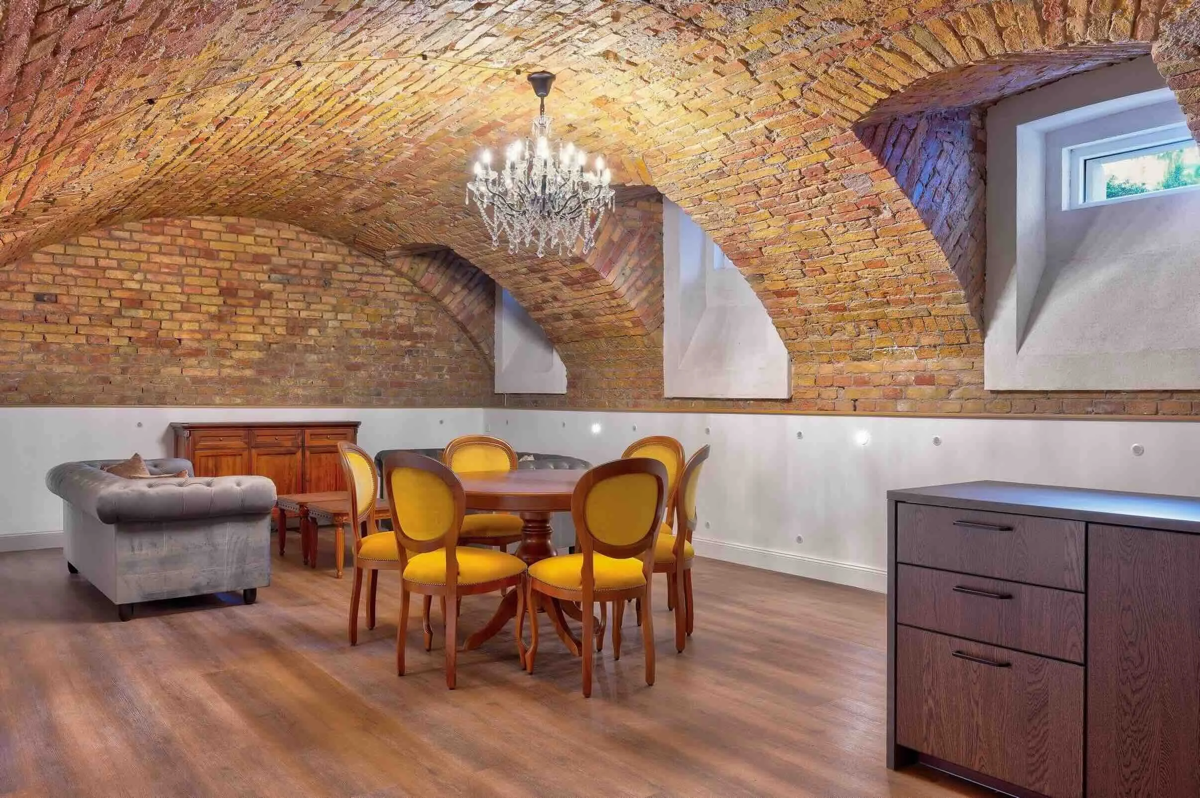 An interior room with an arched brick ceiling, wooden floor, a chandelier, a round dining table with yellow-upholstered chairs, a sofa, a wooden cabinet, and a chest of drawers.
