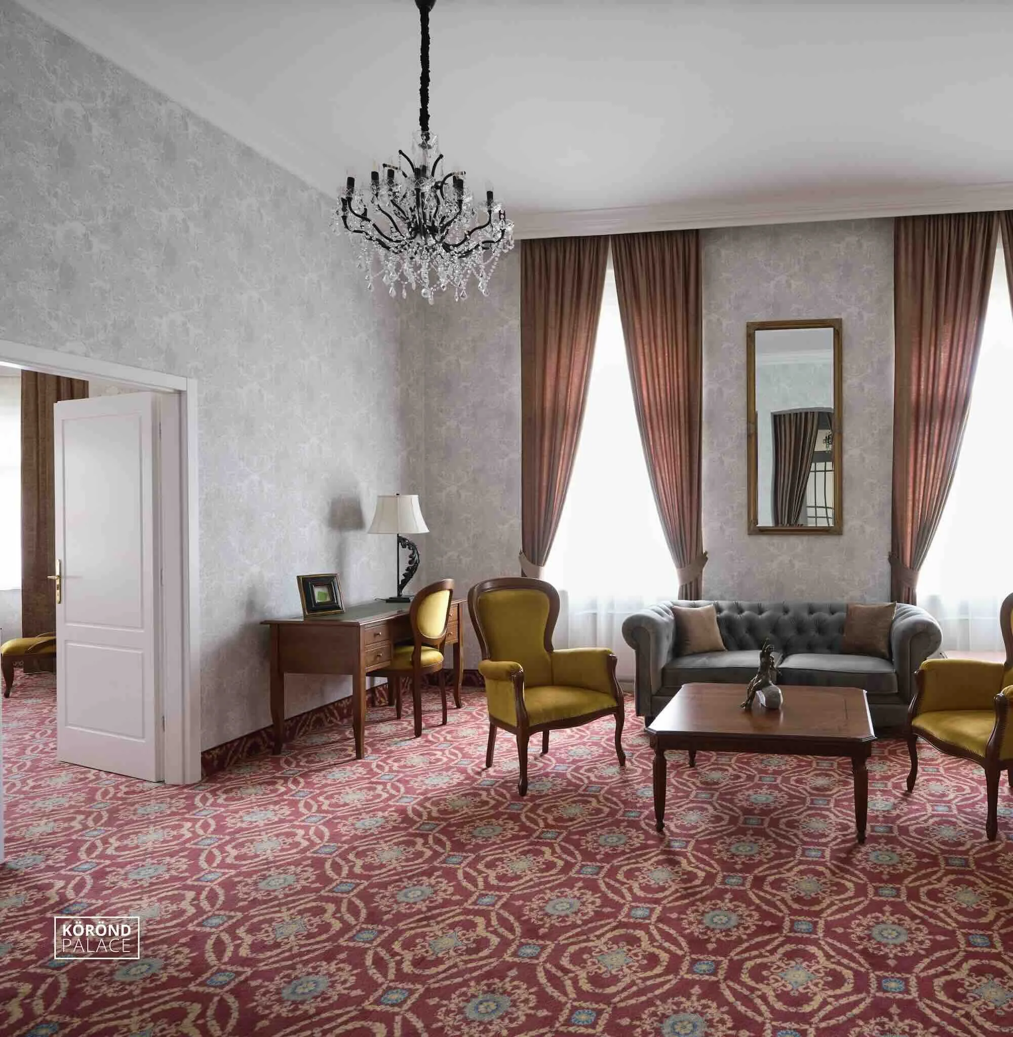 Elegant living room with a chandelier, large windows with red curtains, a gray tufted sofa, yellow armchairs, a wooden coffee table, a desk with a lamp, and a mirror on textured wallpapered walls.