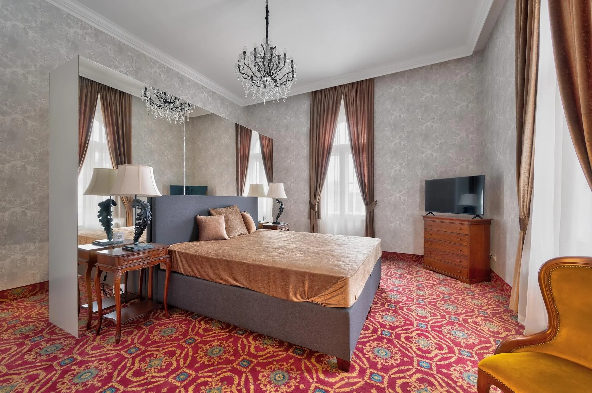 Elegant hotel bedroom with a plush bed, mirrored wall, chandelier, lamps, large windows with brown curtains, a flat-screen TV, and a yellow chair.