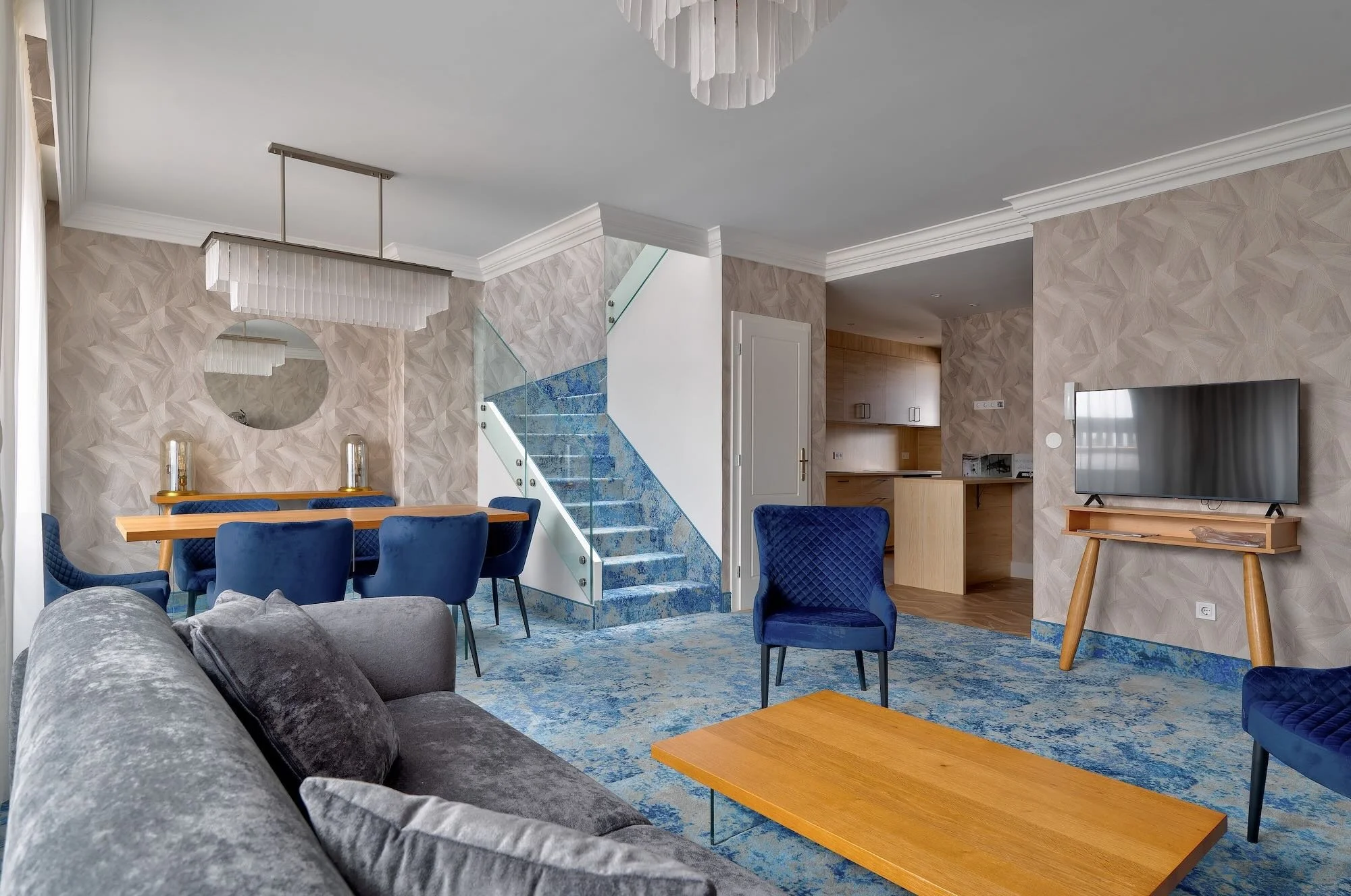 Living room with gray sofa, blue chairs, wooden coffee table, patterned blue carpet, wall-mounted TV, and a staircase with blue floral carpet leading to the upper floor.