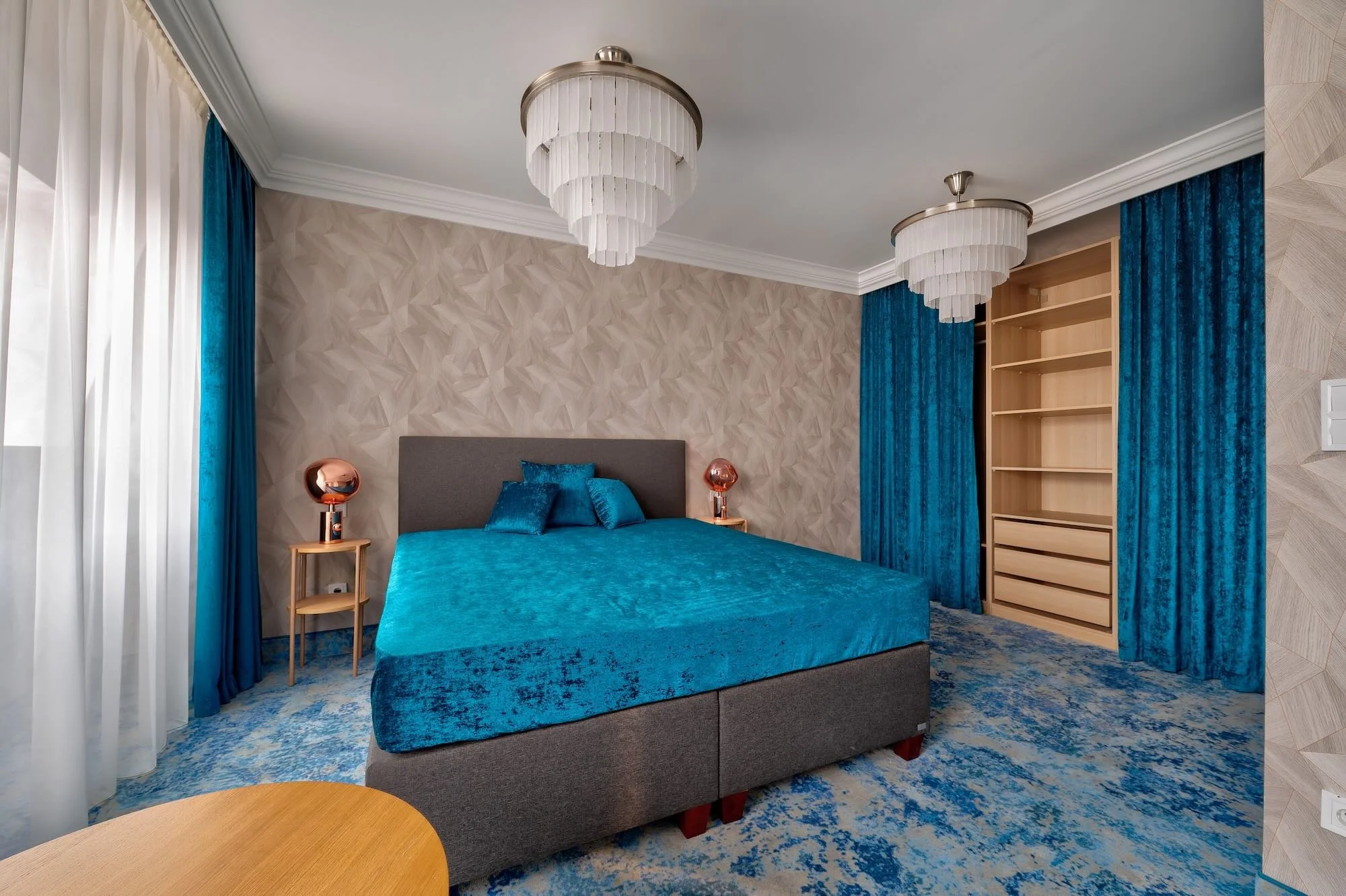Bedroom with grey bed, blue bedspread, blue pillows, beige textured accent wall, floor-to-ceiling blue curtains, wooden side table with copper-colored lamp, empty built-in wooden shelf, two white-tiered chandeliers.