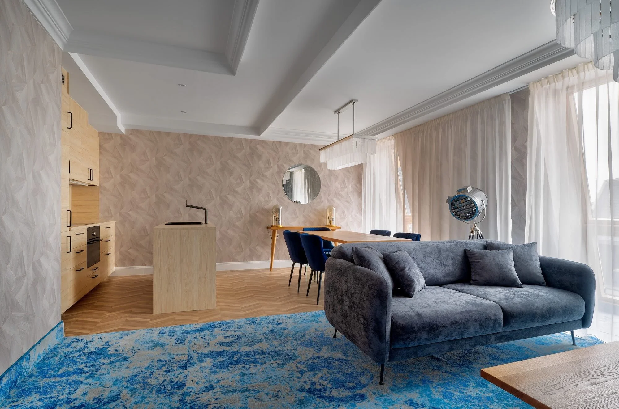 Modern living room with grey velvet sofa, blue area rug, wooden dining table with six blue chairs, open kitchen with light wood cabinets, geometric textured wall, large windows with sheer white curtains, circular mirror on the wall, and a large film 