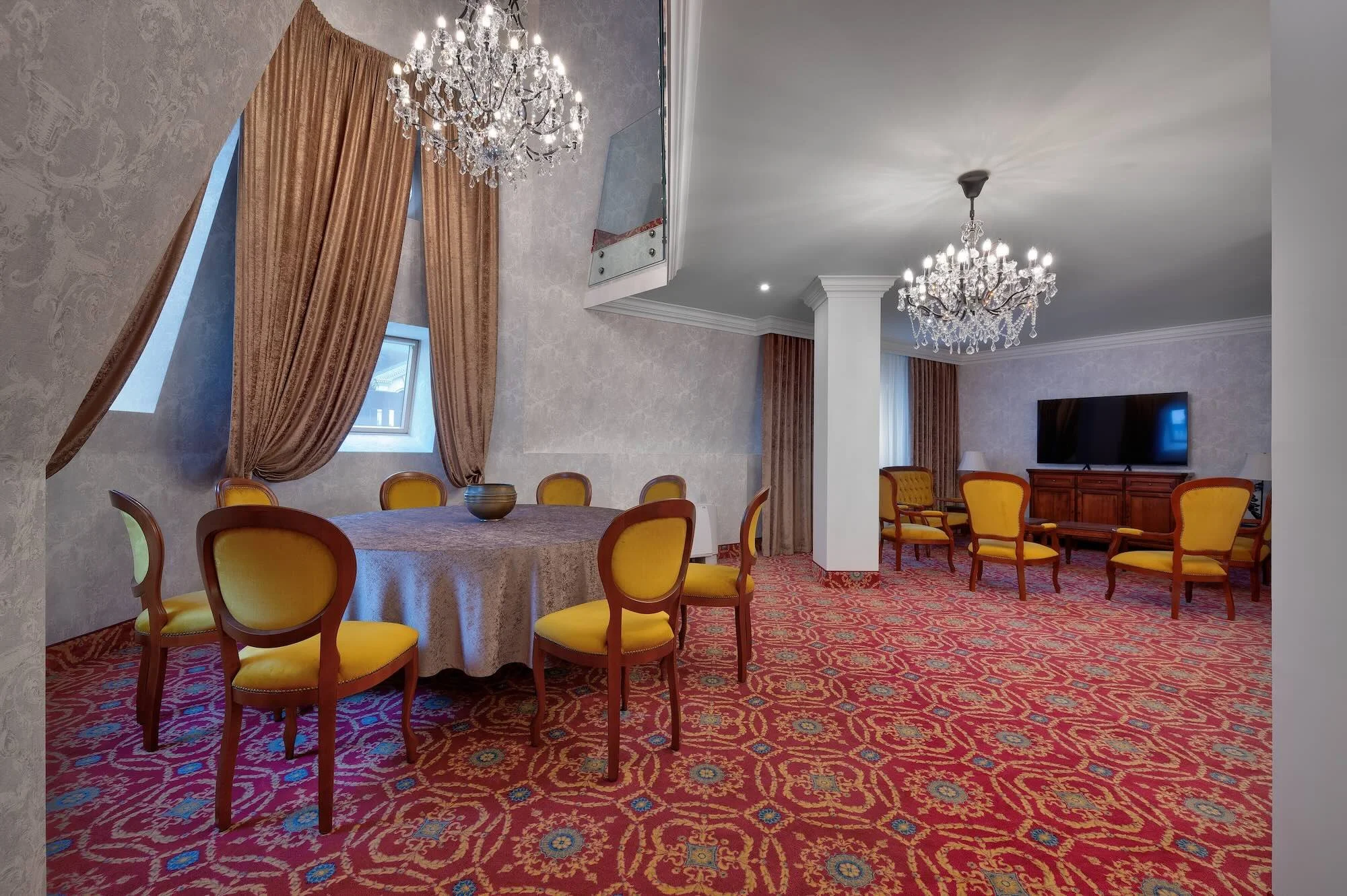 Elegant room with two chandeliers, a round table with yellow chairs and a bowl, and a sitting area with yellow chairs, a wooden cabinet, and a large flat-screen TV on the wall.