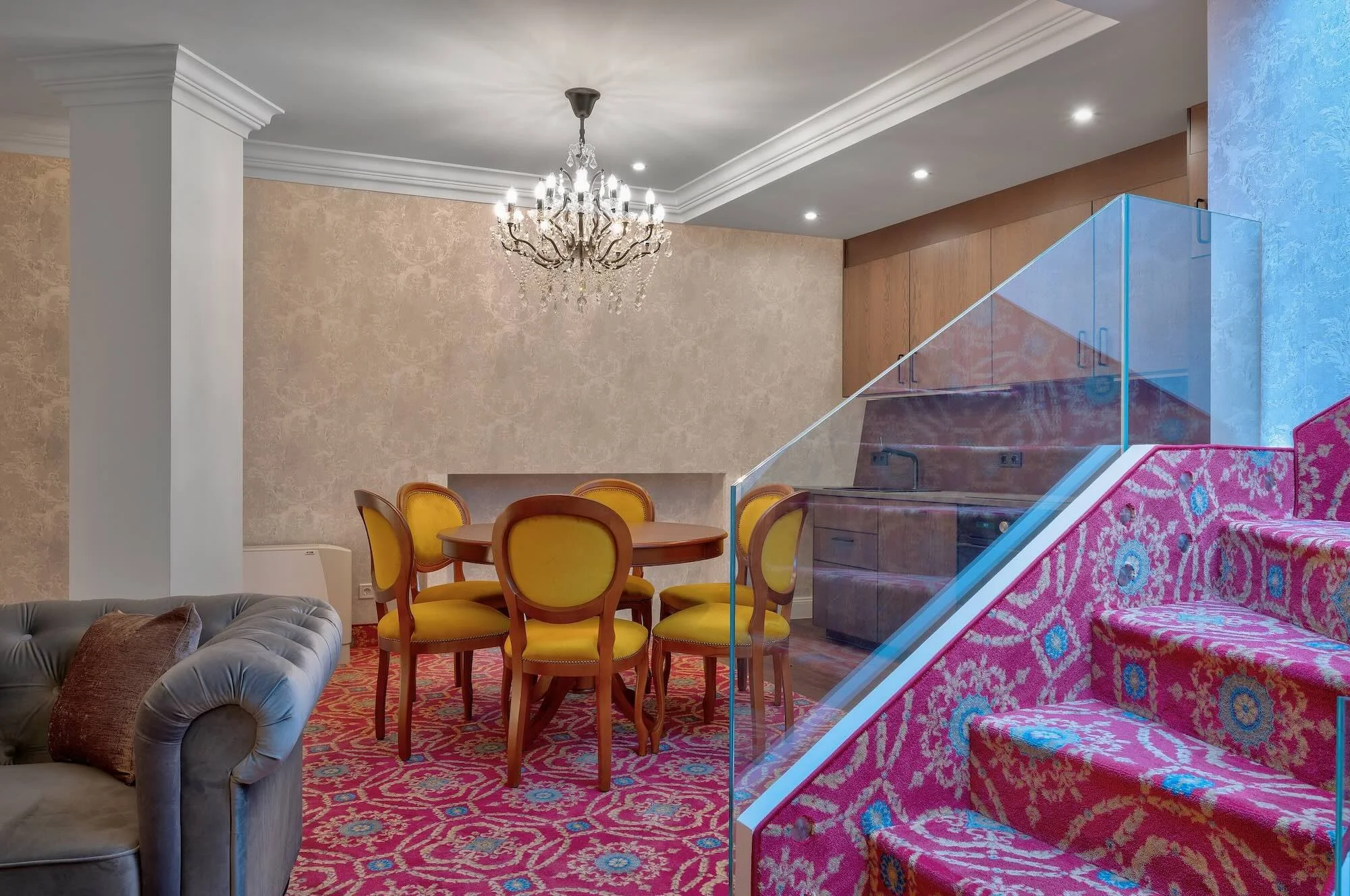 Living room with beige textured walls, a chandelier, yellow dining chairs around a round wooden table, a gray sofa with a brown pillow, a pink patterned staircase with a clear glass railing, and a kitchen area with wooden cabinets.