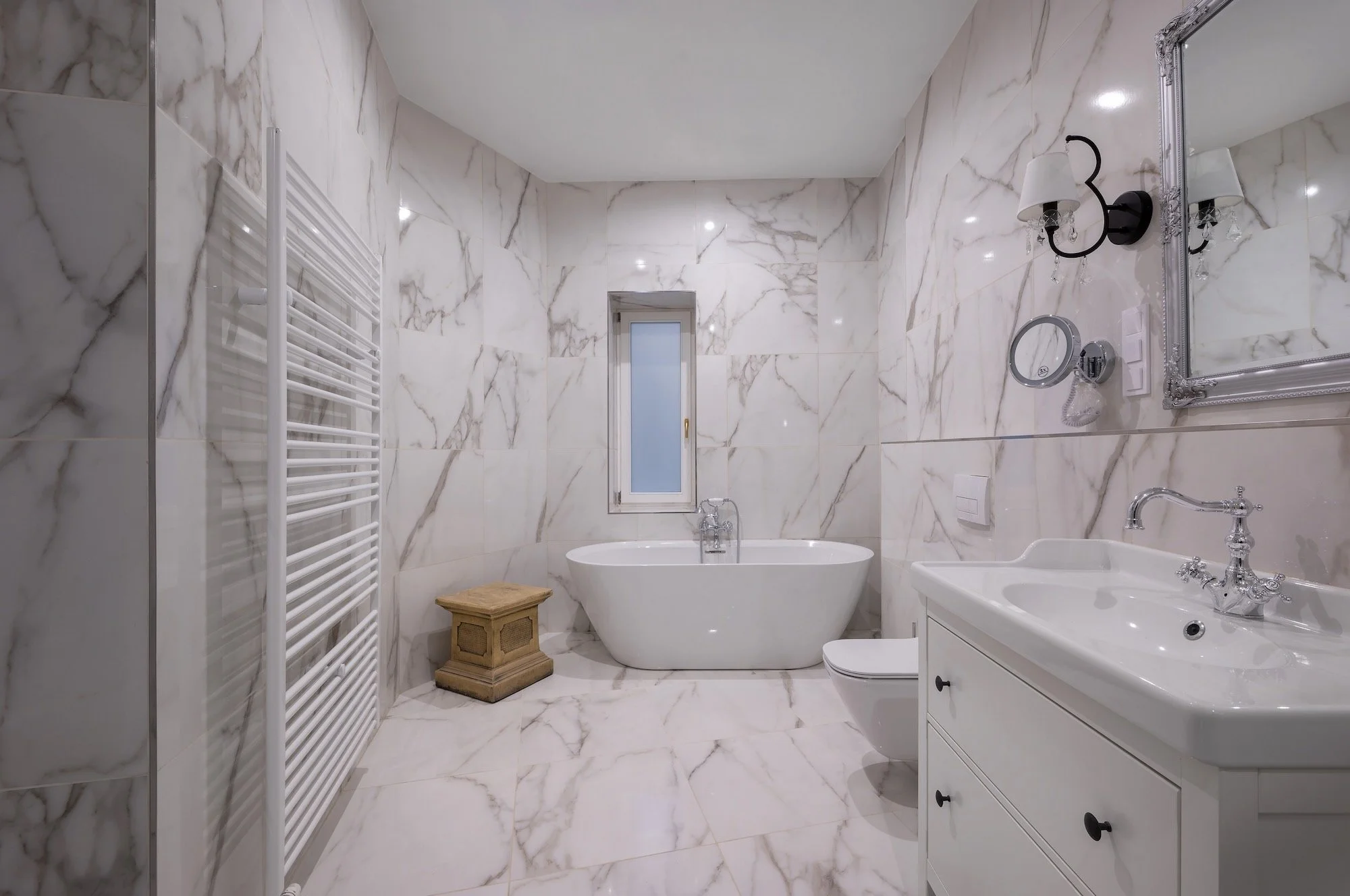 Bathroom with marble walls and floor, featuring a freestanding bathtub, a small window, a white sink vanity with a mirror, and a wall-mounted light fixture.