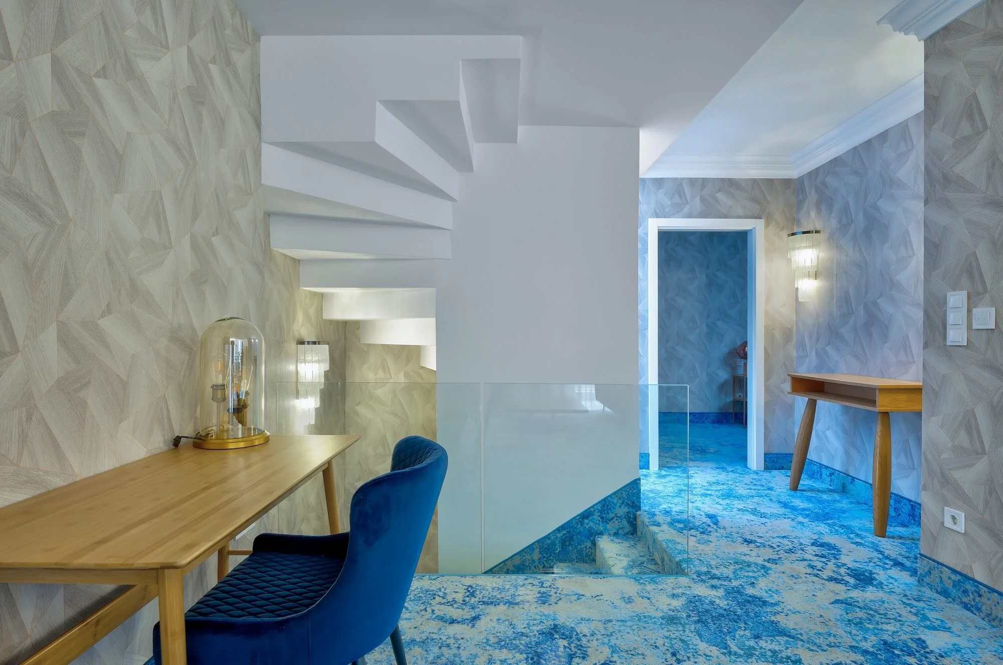Interior view of a modern home with a staircase, a wooden desk with a blue chair, textured wallpaper, blue patterned carpet, and a doorway.