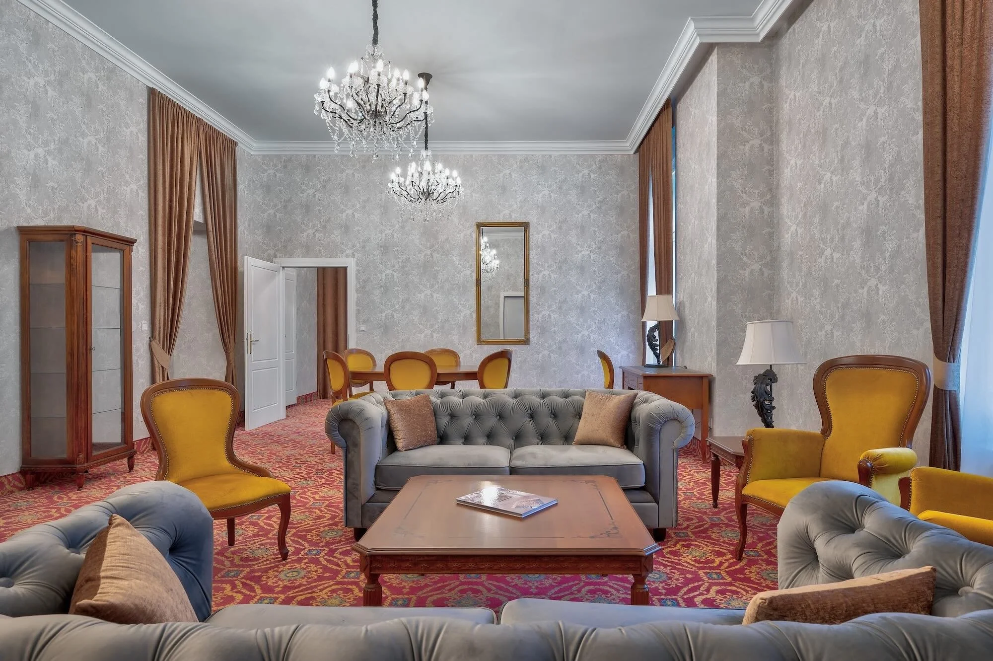 Elegant hotel lounge with gray and yellow armchairs, a gray tufted sofa, a wooden coffee table, gold accents, chandeliers, large windows with brown curtains, and framed mirrors.