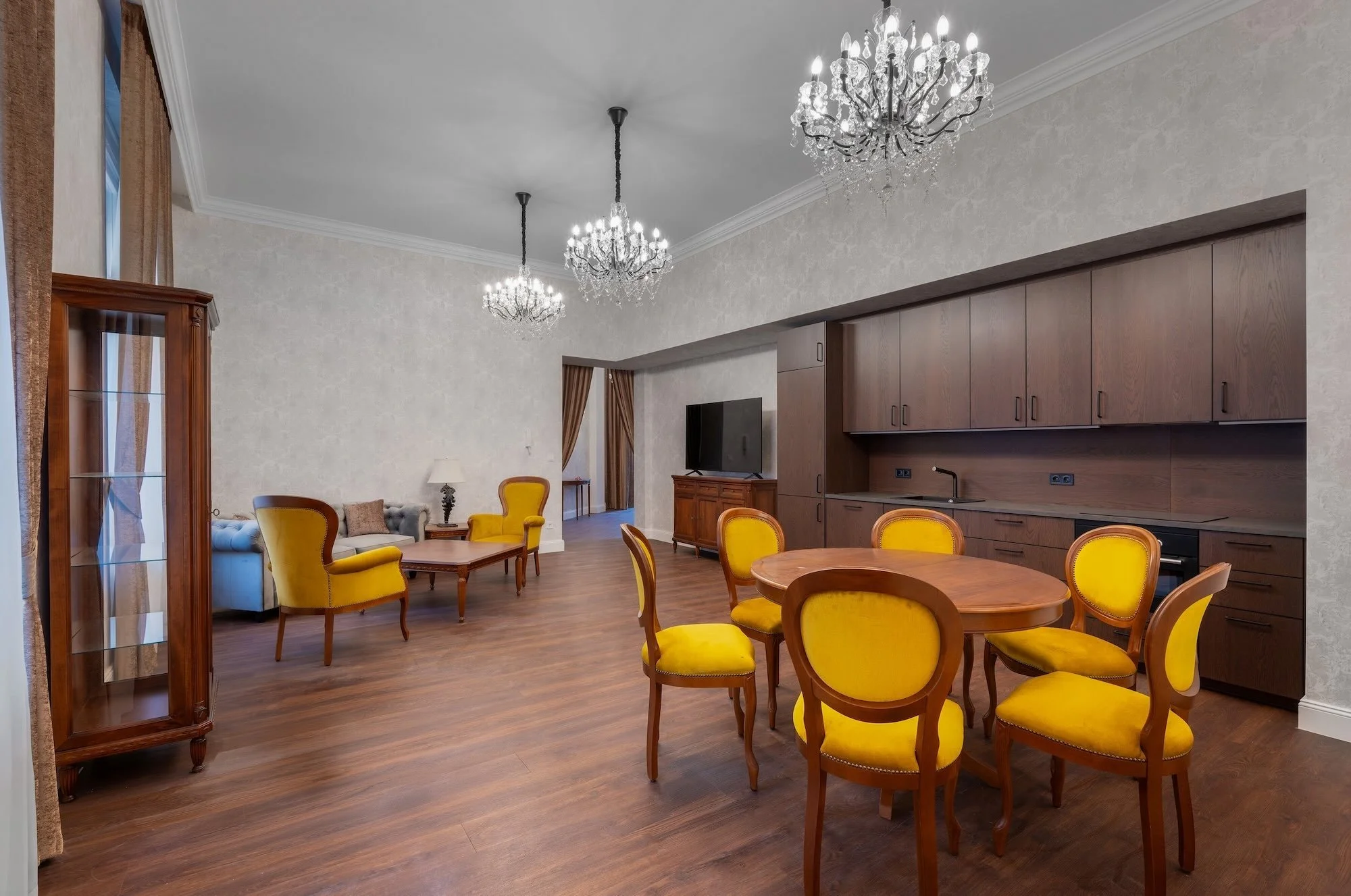 Living and dining room with wooden flooring, yellow upholstered chairs around a wooden dining table, a wooden display cabinet, chandeliers, television, and sofa with curtains in background.