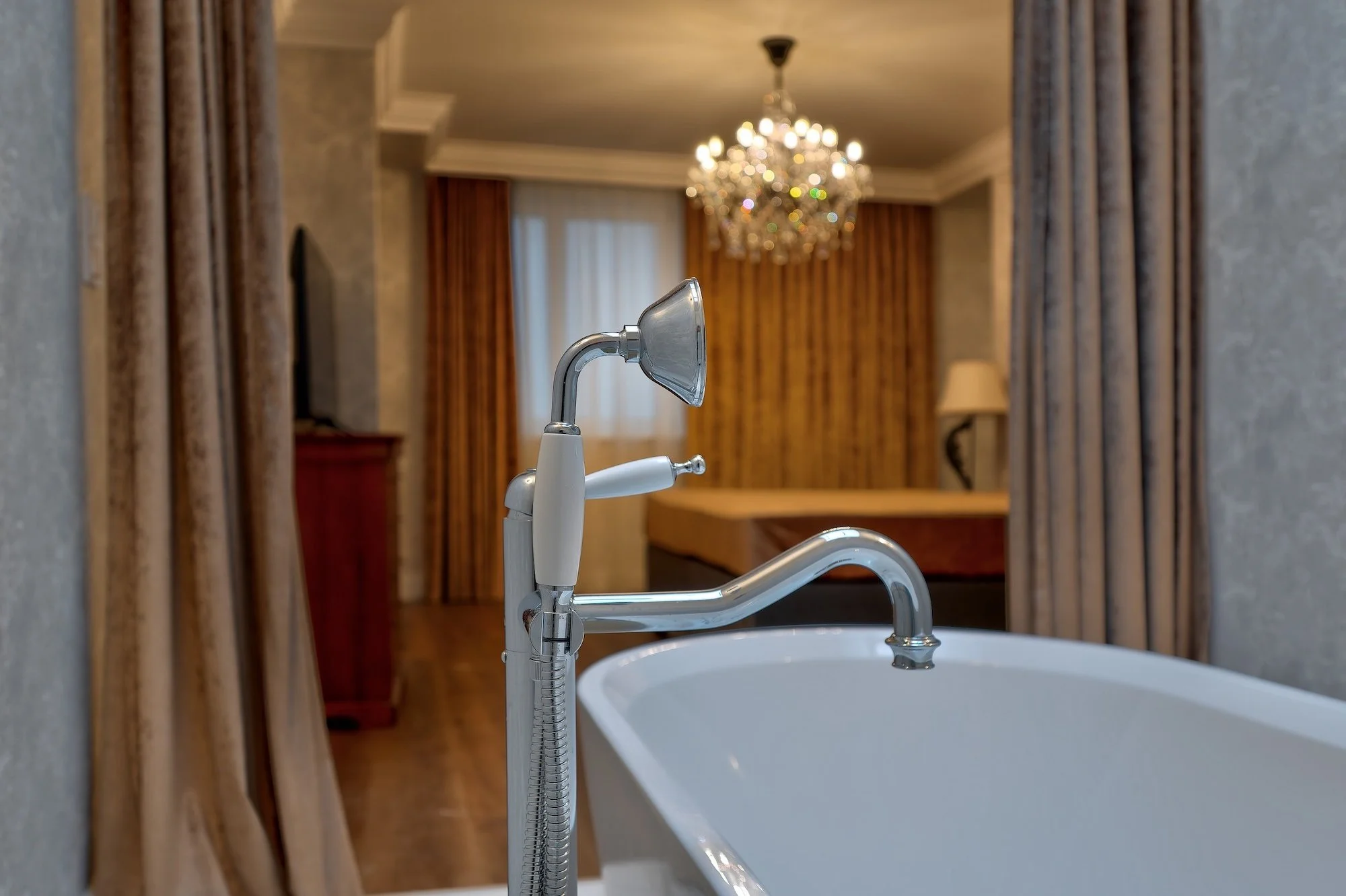 Bathtub with chrome faucet and handheld shower head in a luxurious bathroom with gold and beige curtains, a chandelier, and a wooden dresser in the background.