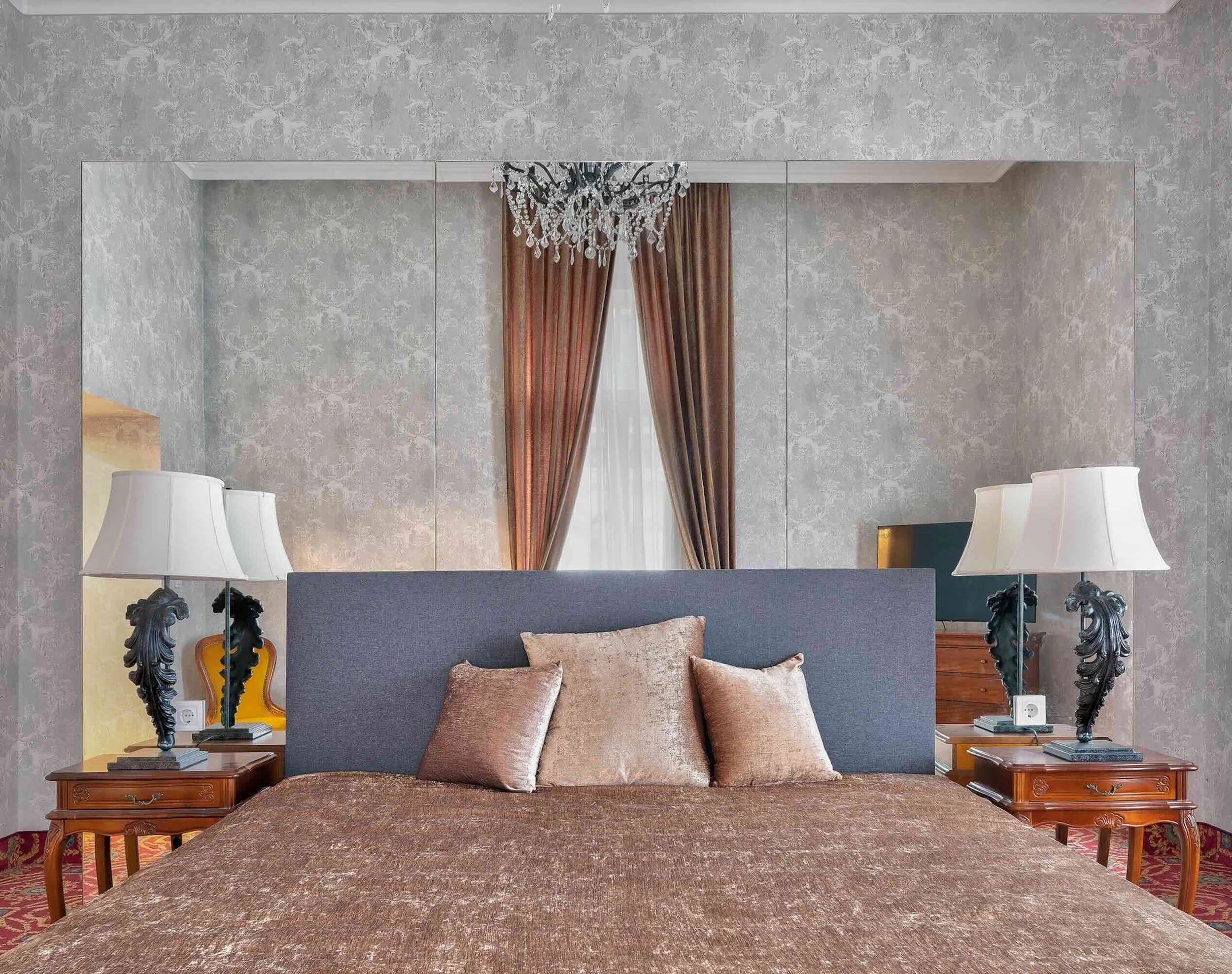 A luxury hotel bedroom with a large bed, three beige throw pillows, mirrored wall, chandelier, and side tables with lamps.