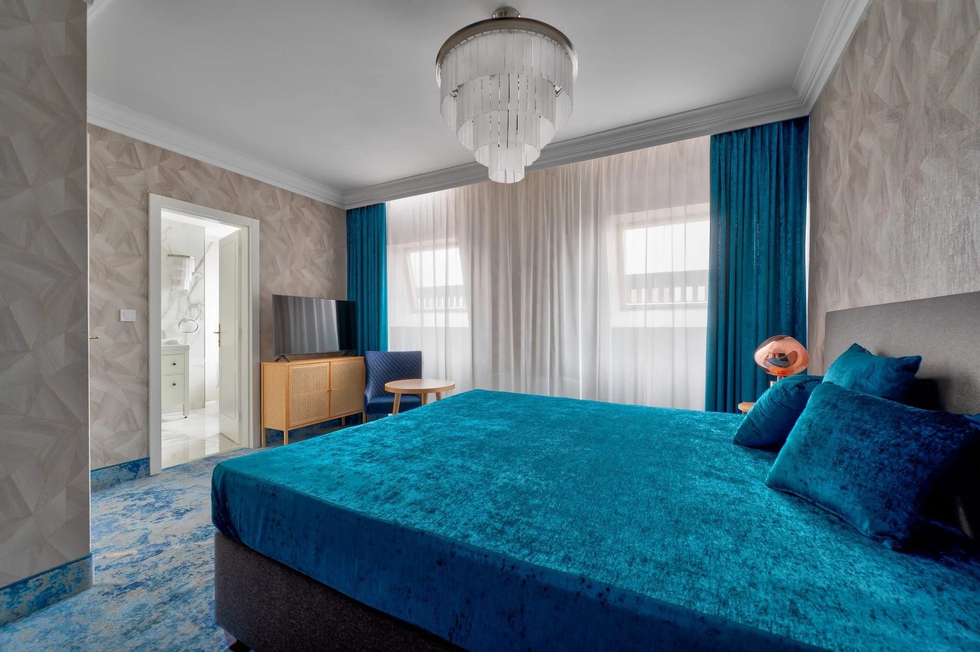 A hotel bedroom with a bed covered in blue velvet, matching pillows, and a gray headboard. There is a blue curtain and sheer white drapes on the window. A wooden bedside table with a pink lamp, a small wooden table, a TV on a wooden stand, and a blue