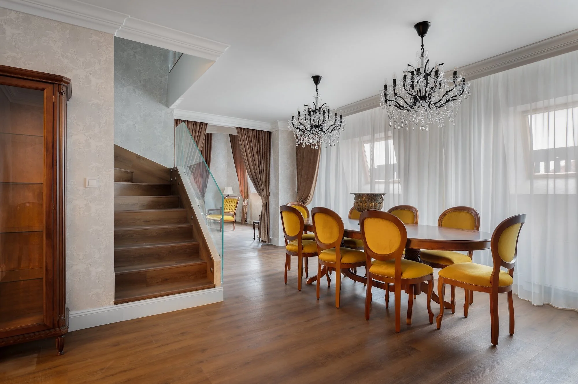 Elegant dining room with a large wooden table surrounded by yellow upholstered chairs, chandeliers hanging from the ceiling, large windows with sheer curtains, and hardwood flooring.
