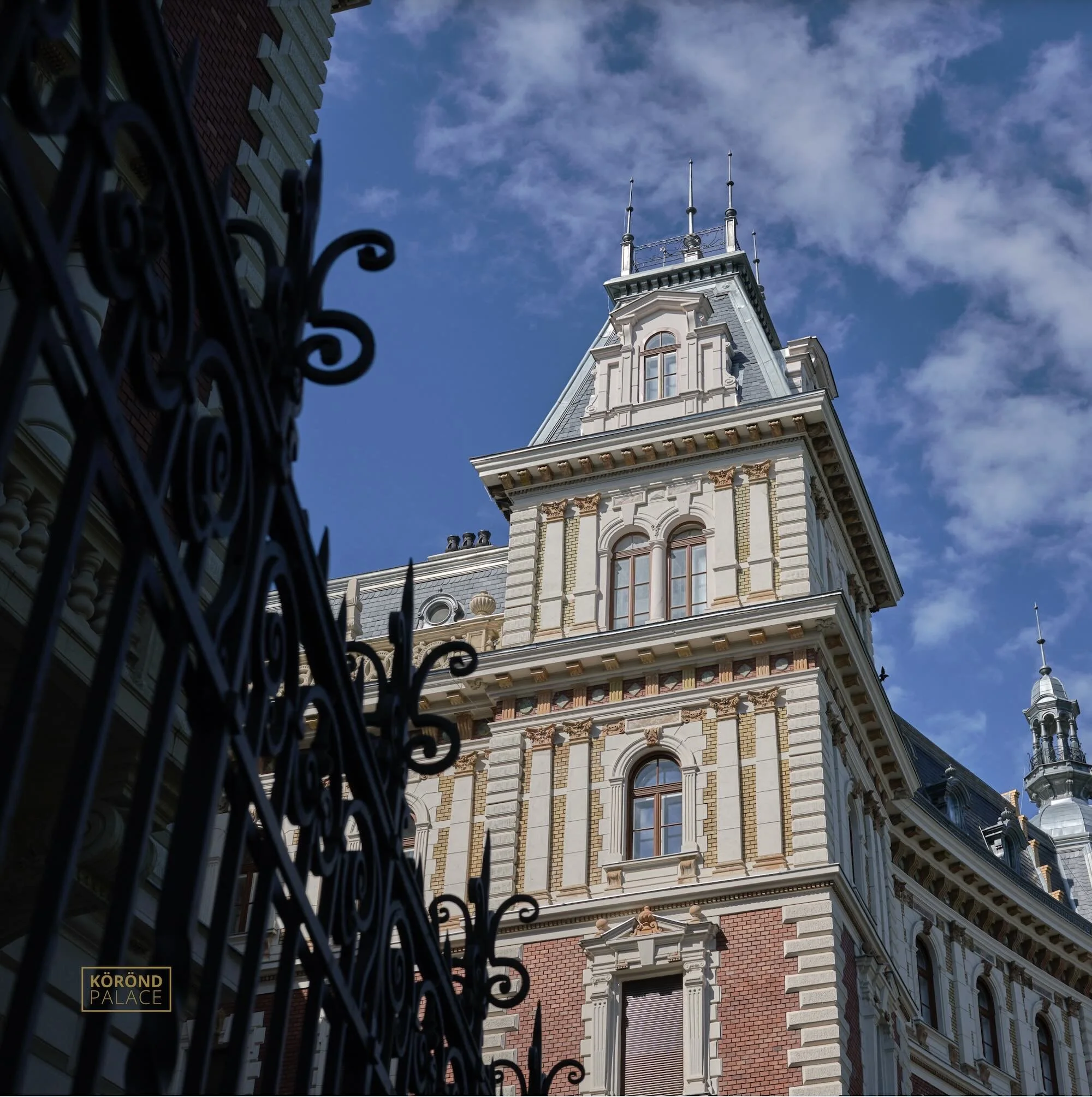 A historic European-style building with ornate architectural details, tall windows, and a pointed tower, partially obscured by a black wrought iron fence in the foreground, under a partly cloudy blue sky.