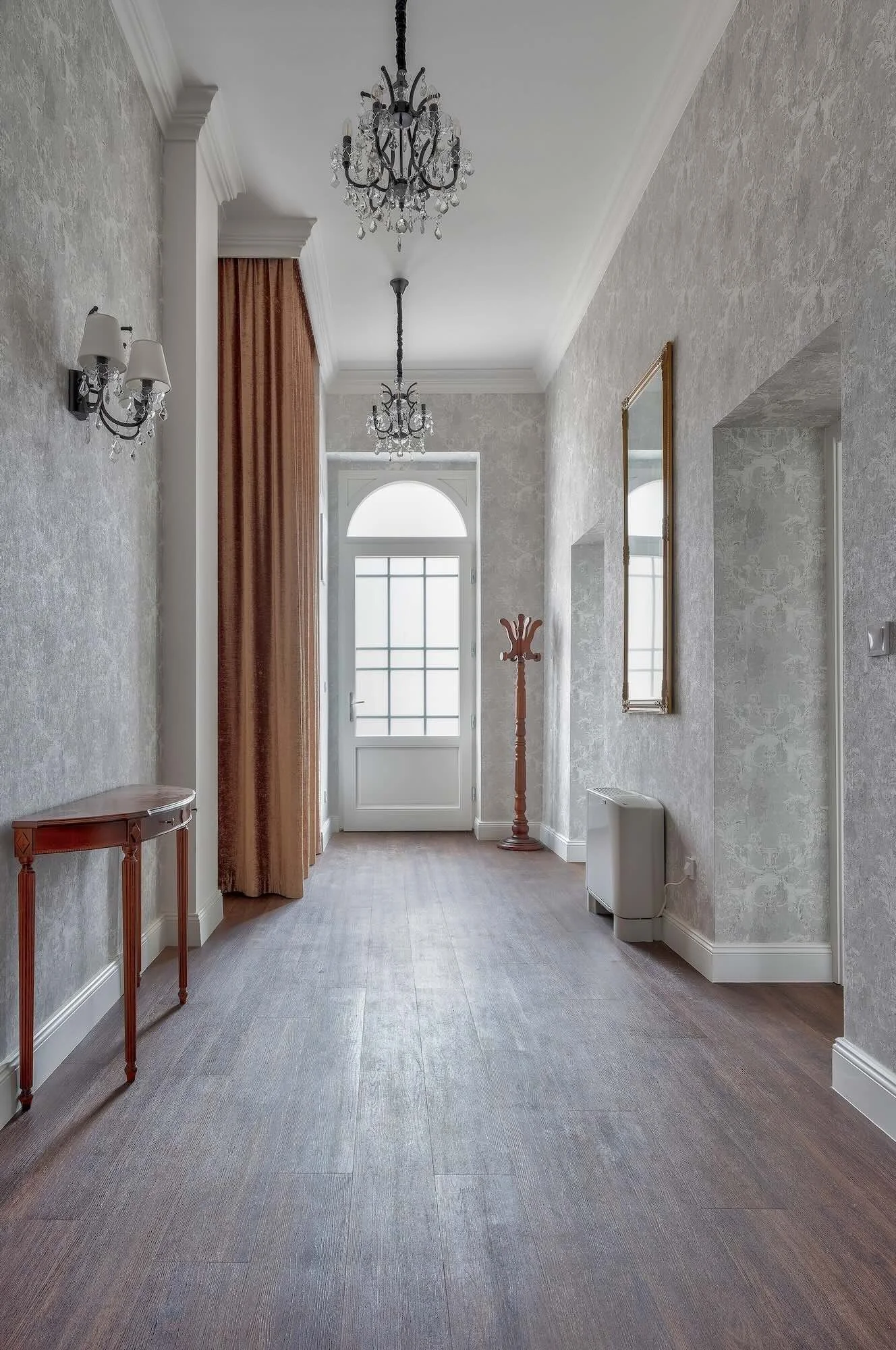 Interior hallway with chandelier lighting, floral patterned wallpaper, a small wooden table on the left, a mirror on the right wall, and a coat stand near the door at the end of the hallway.