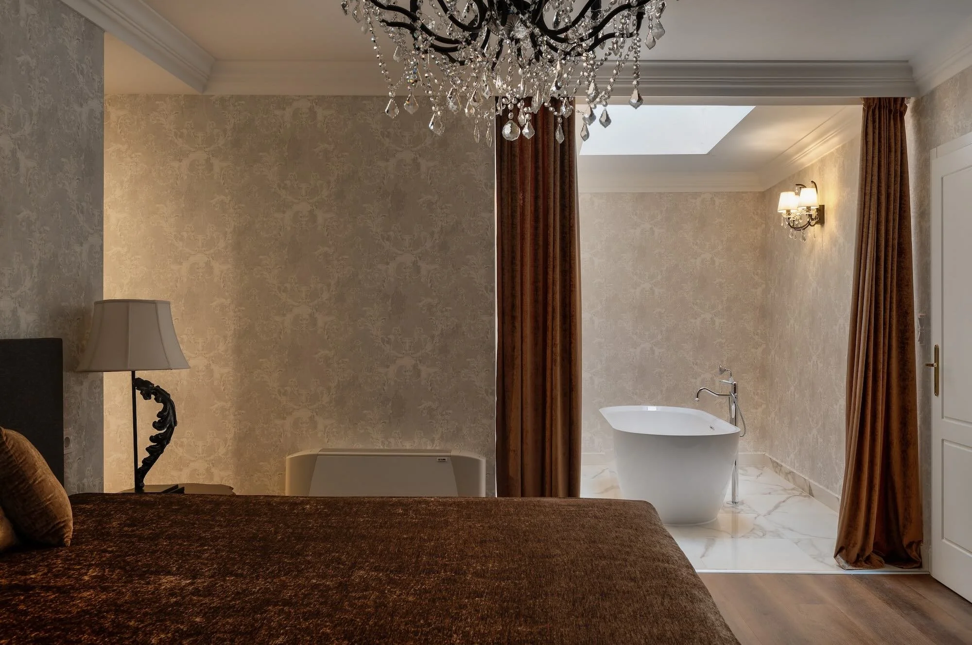 A bedroom with a large bed covered in brown bedding, a nightstand with a beige lamp, and a chandelier hanging from the ceiling. Behind a curtain, there is a bathroom area with a free-standing bathtub, a skylight, and wall-mounted lights.
