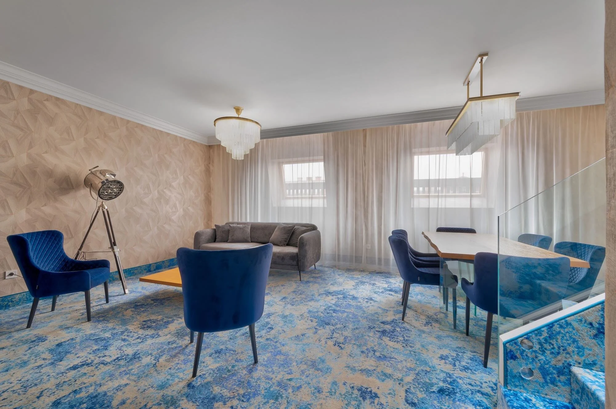 Living room with blue velvet chairs, gray sofa, patterned wallpaper, chandelier, and large windows with sheer curtains.