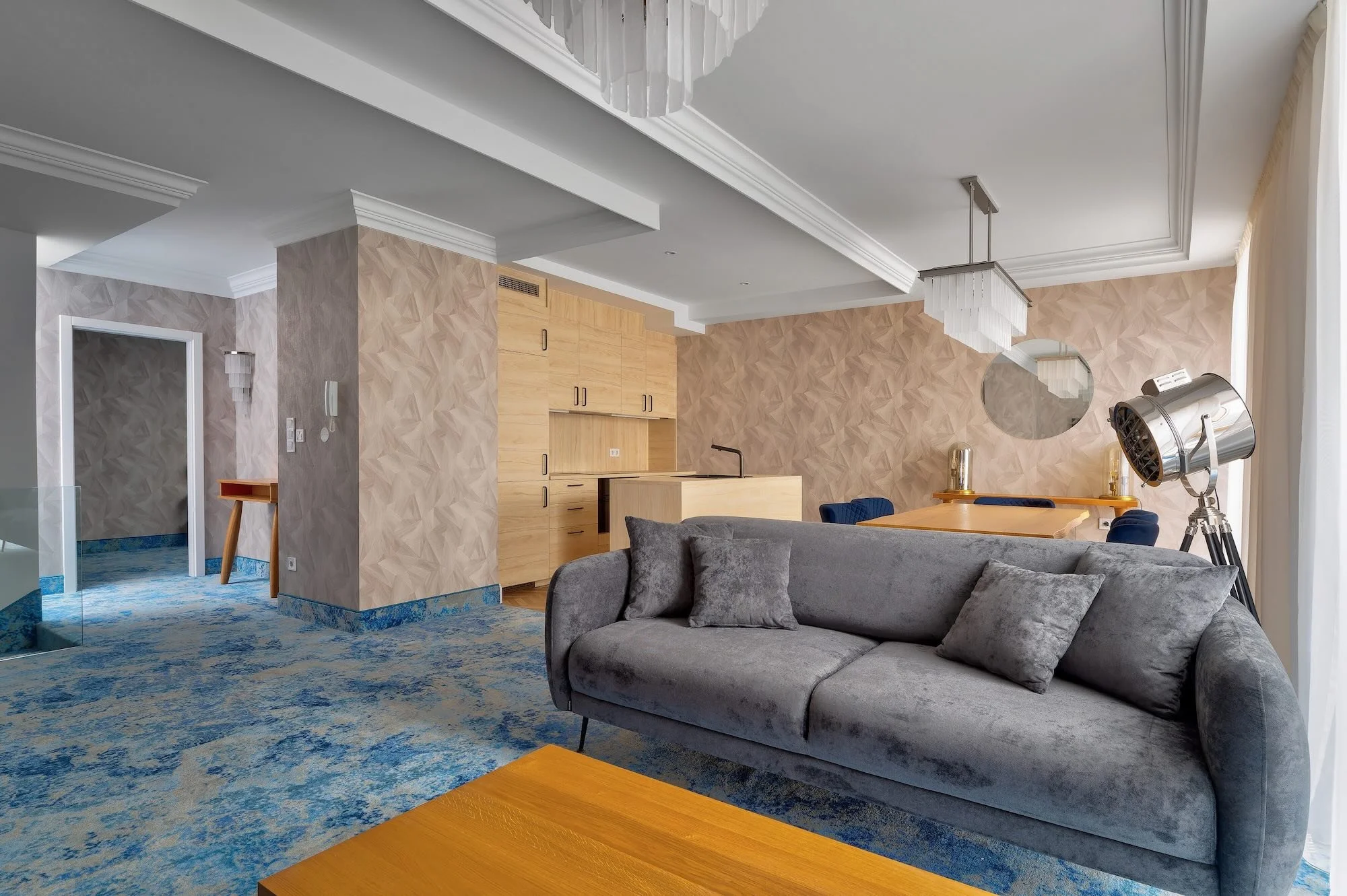 Living room with gray velvet sofa, wooden coffee table, blue and gold accents, beige textured walls, and a modern chandelier. There's an open kitchen with wooden cabinets and a dining area with a wooden table and blue chairs. A large floor lamp and c