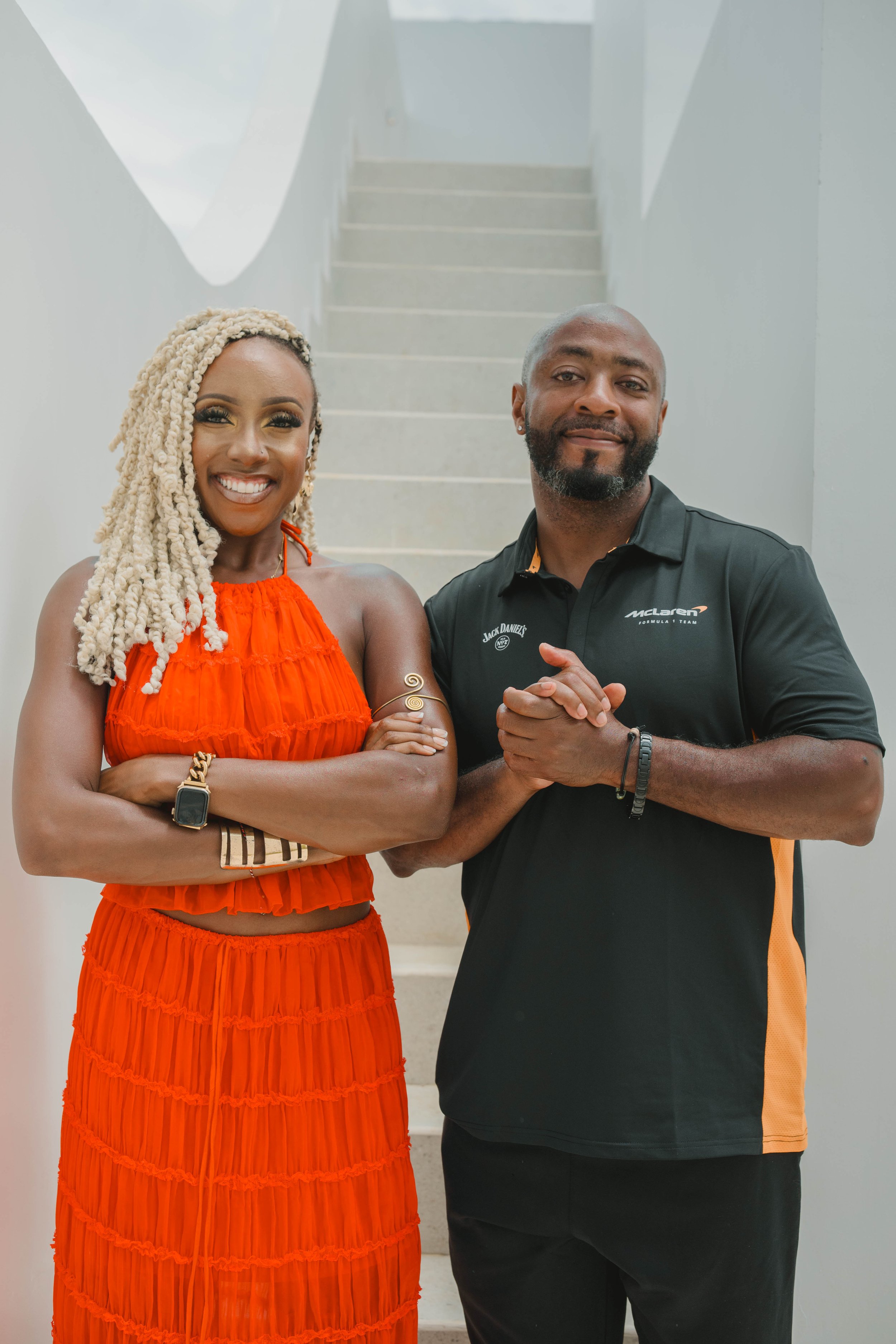 A smiling woman in a bright orange dress and a man in a black McLaren shirt standing together on a staircase. Both have their arms crossed or clasped, the woman has blonde dreadlocks, and the man has a beard.