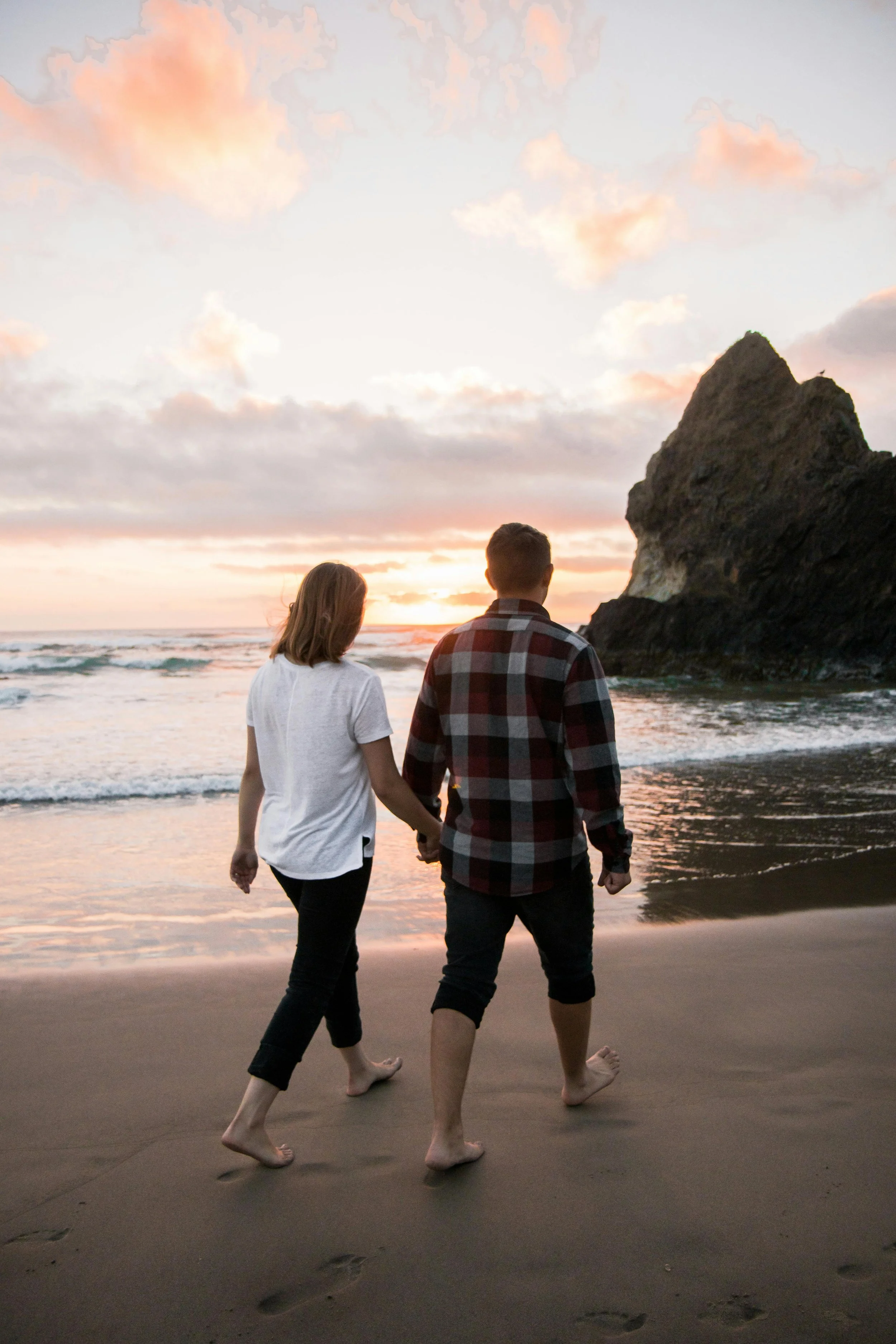 A couple walking hand in hand along the beach at sunset, with a large rocky formation in the background.