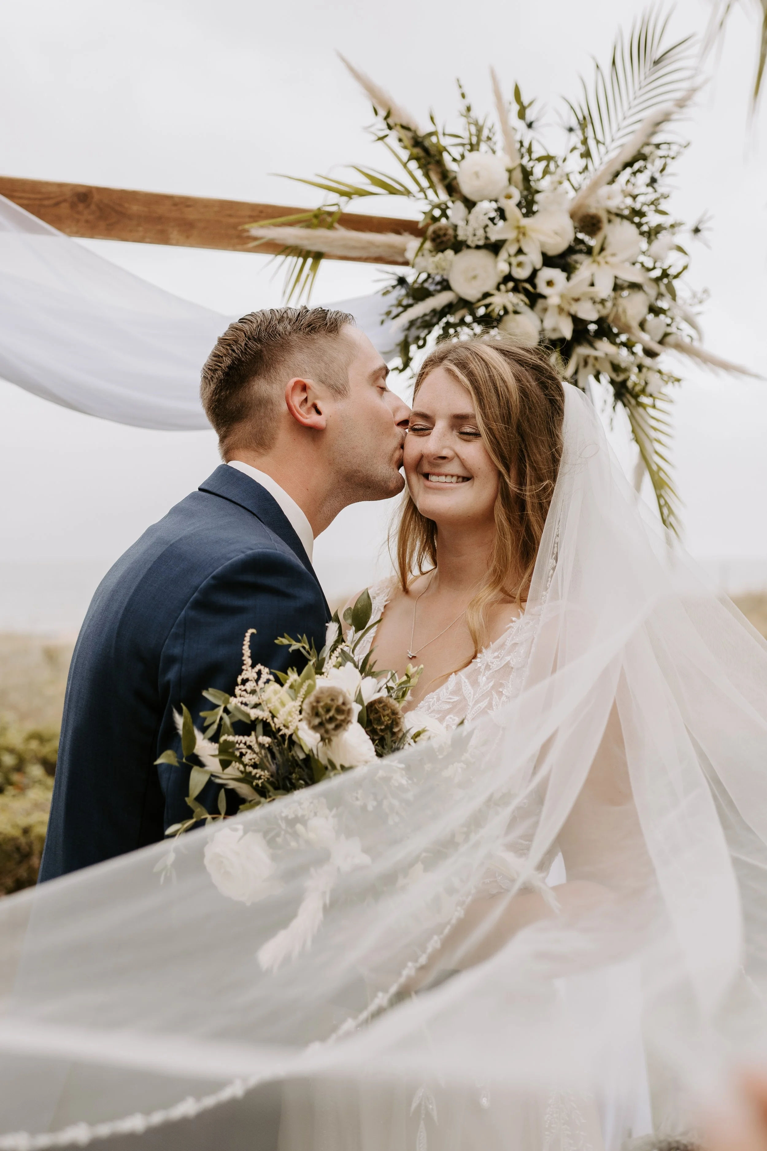 A newly married couple shares a moment under a wedding arch decorated with white flowers and greenery, with the groom kissing the bride's cheek as she smiles.