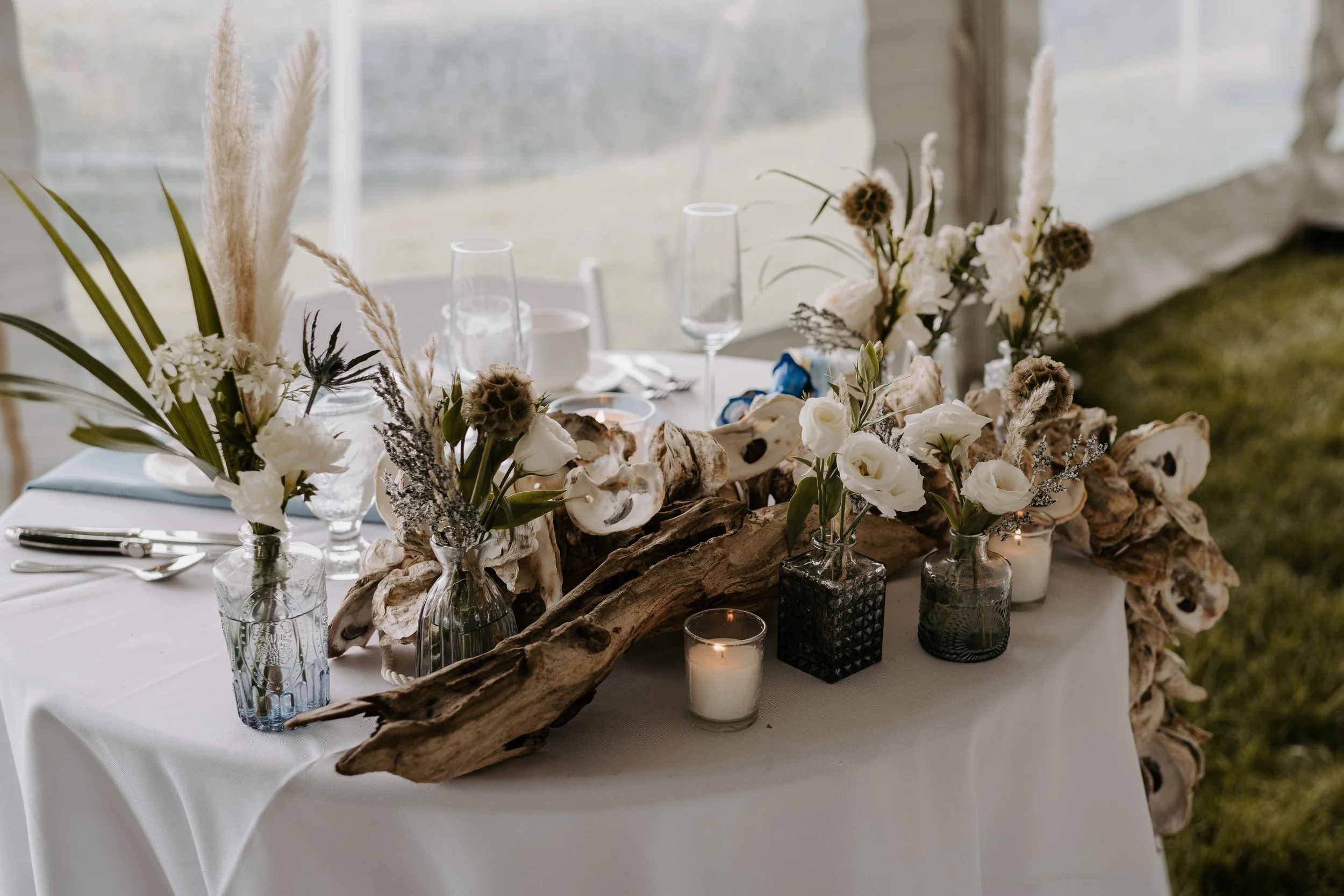 Elegant table centerpiece with white flowers, dried seed pods, candles, and driftwood on a white-draped table.