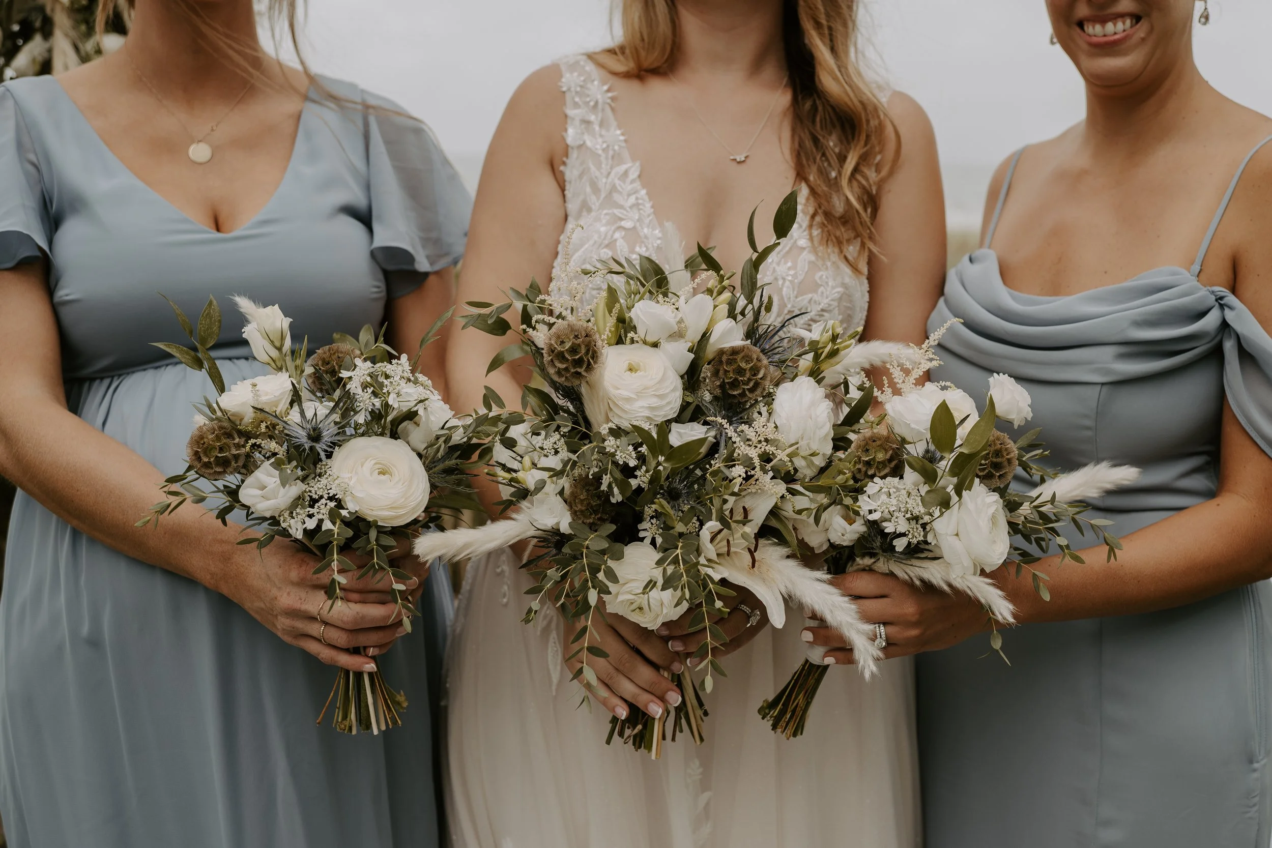 Close-up of three women, a bride in the center and two bridesmaids, holding bouquets of white flowers and greenery
