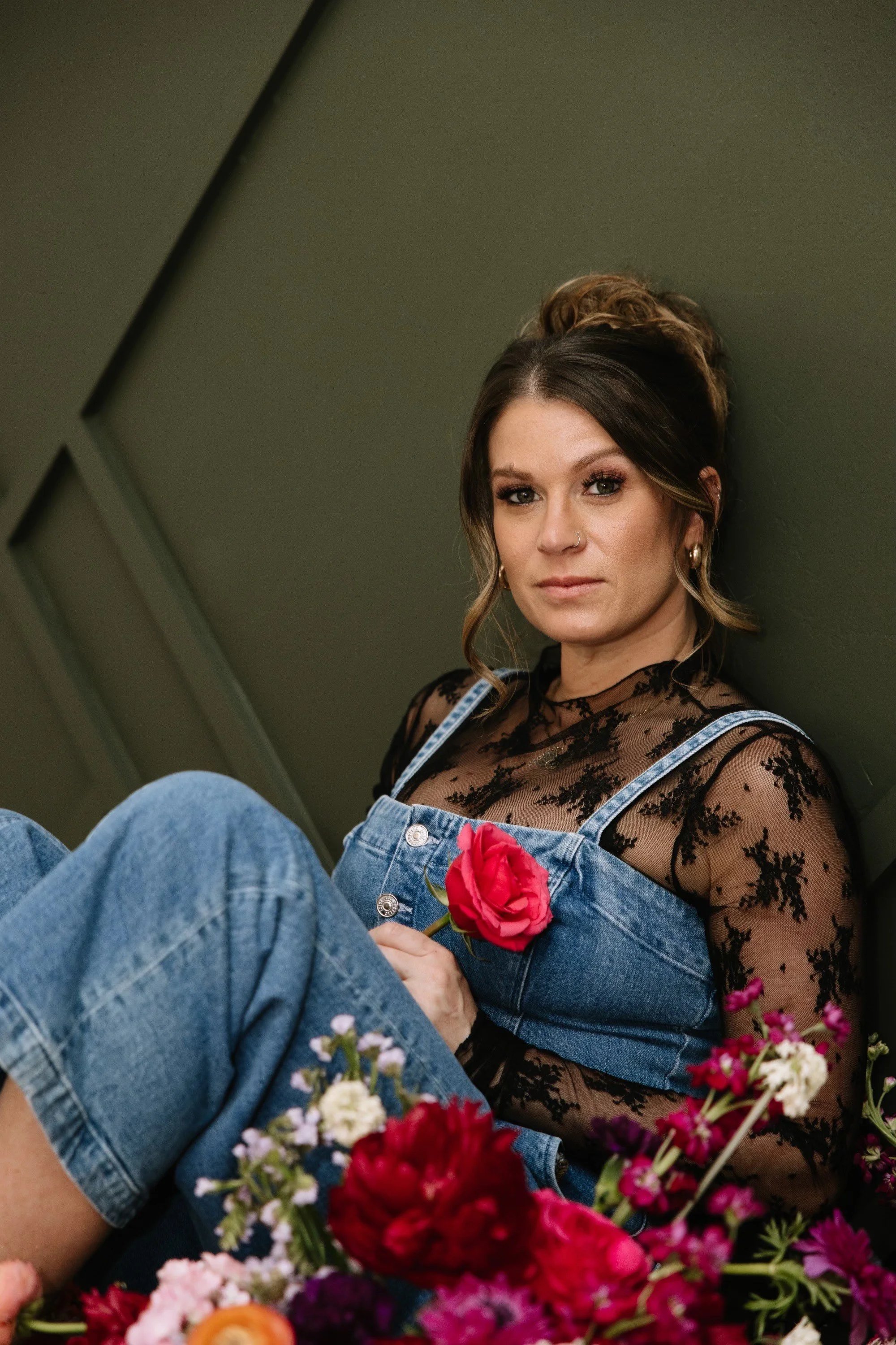A woman with brown hair in a bun, wearing a black lace top under denim overalls, sitting against a green wall, holding a red rose, surrounded by colorful flowers.