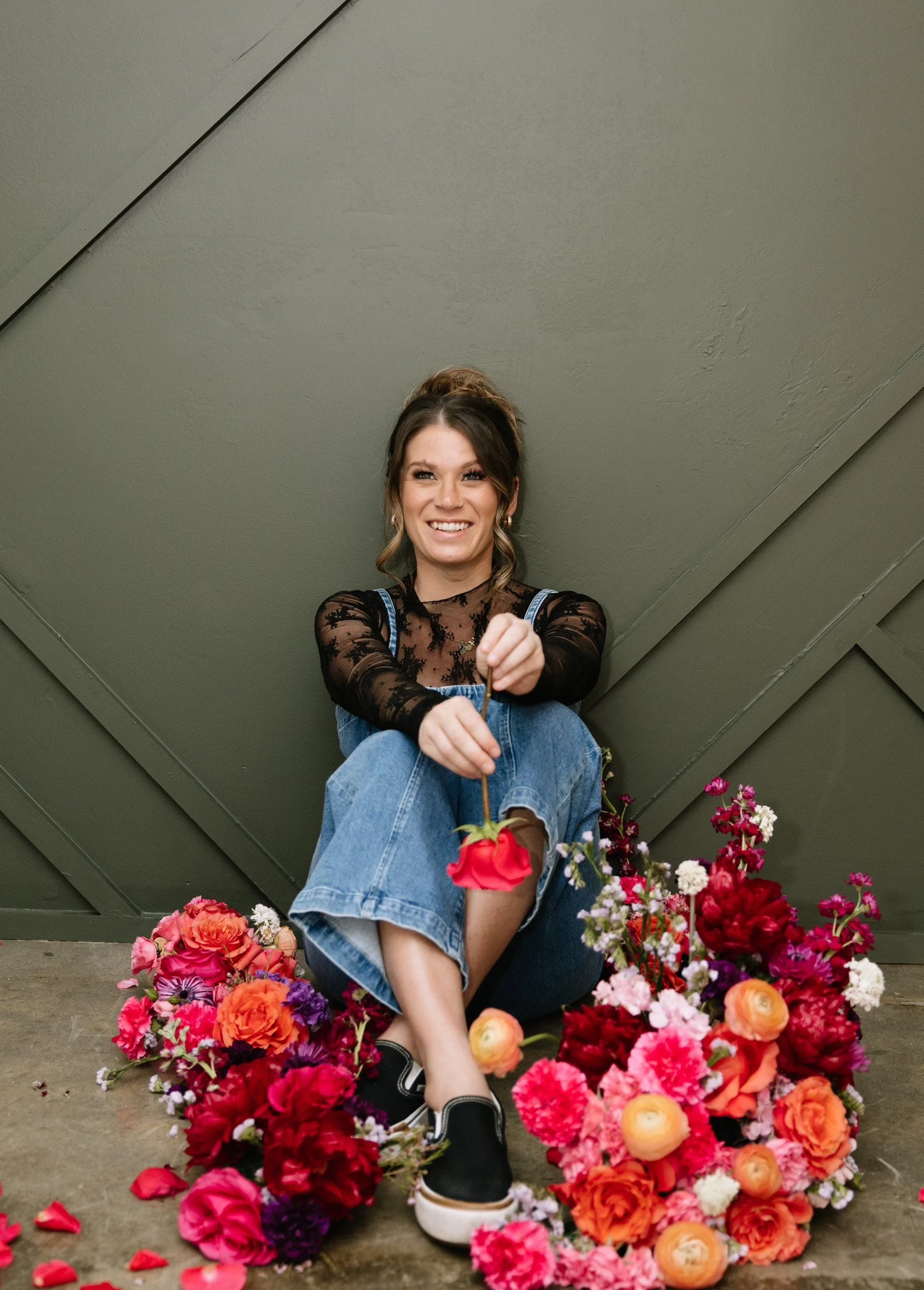 A young woman with brown hair and a black lace top sitting on the floor surrounded by pink, red, purple, and white flowers, holding a pink flower in her hand, smiling at the camera against a greenish-gray wall.