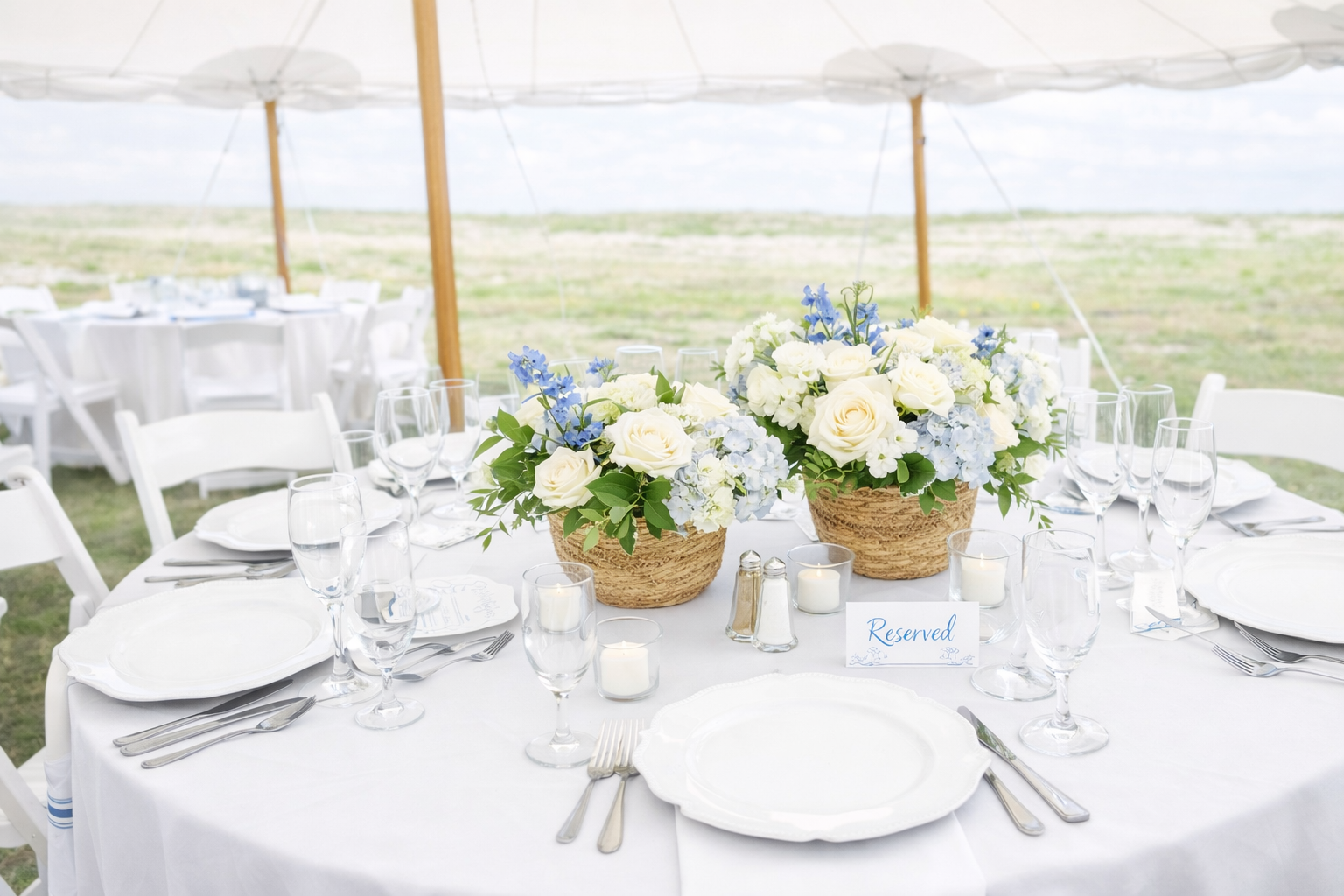 Wedding reception table decorated with white flowers in baskets, surrounded by wine glasses, candles, plates, and silverware, with a 'Reserved' sign, under a tent in an outdoor setting.
