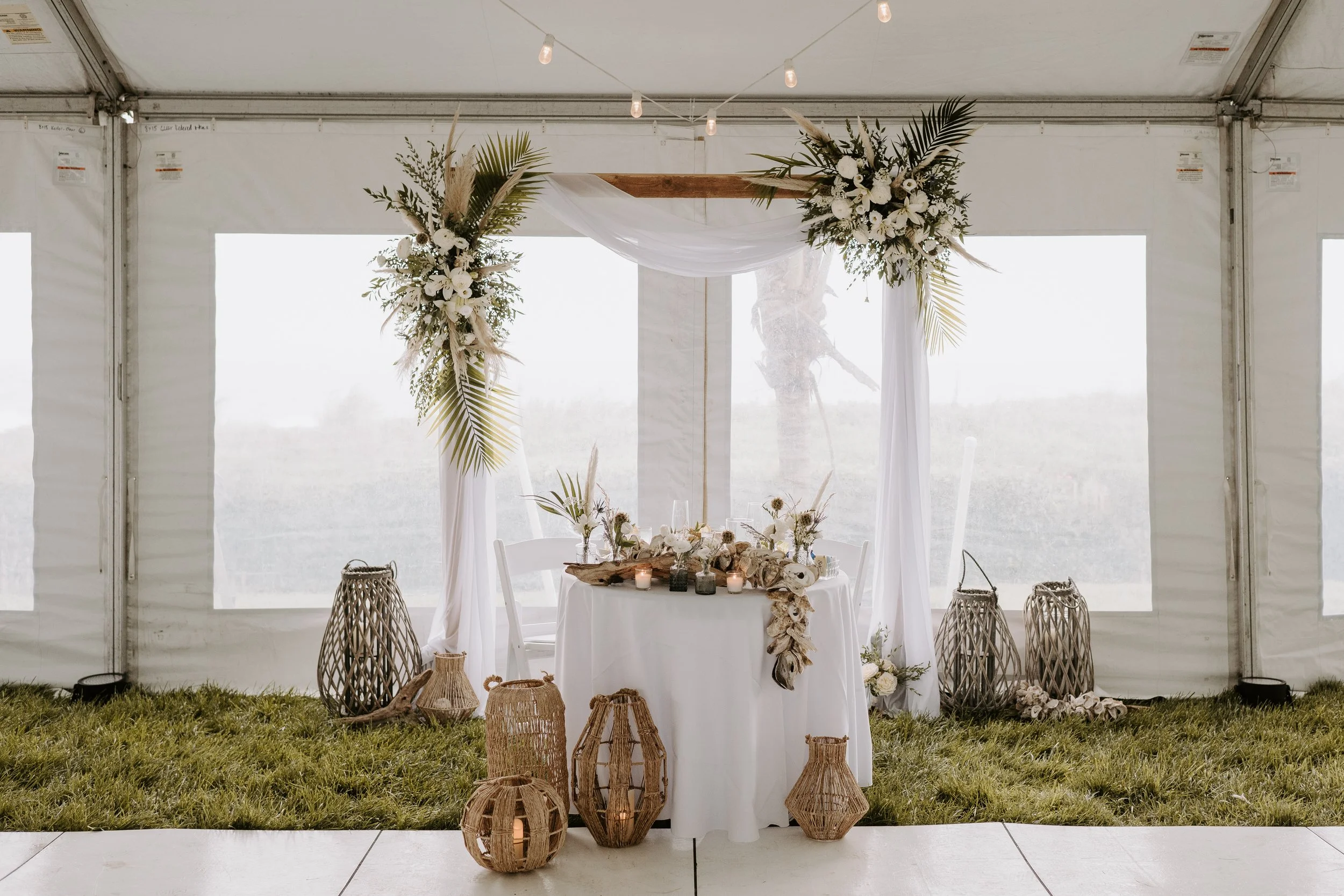 Elegant wedding ceremony setup inside a white tent with a floral decorated arch, a round table adorned with candles, flowers, and natural accents, surrounded by woven lanterns and greenery on grass.
