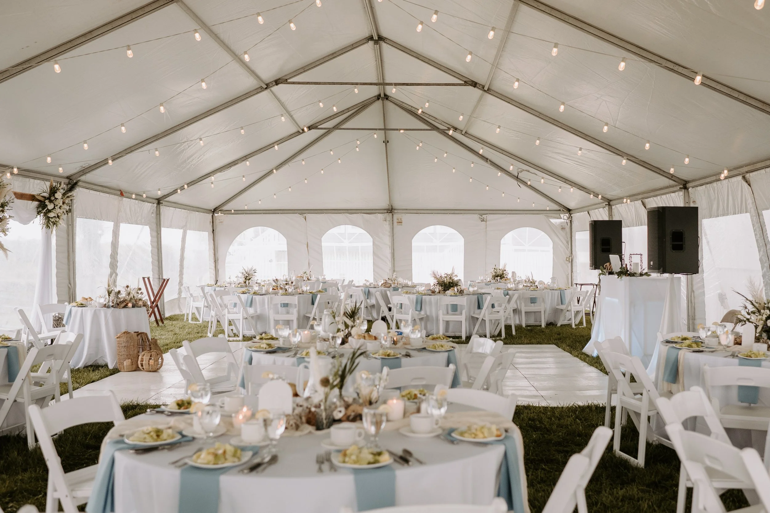Inside a decorated event tent with round tables set for a celebration, featuring white chairs, floral centerpieces, candles, and a DJ setup with speakers, under string lights.