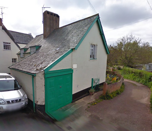 A small house with a steep gable roof, white walls, and a bright green garage door, located next to a parking area with a silver car and a dirt path leading around the house.