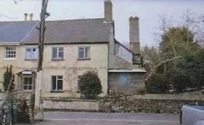 A building with multiple windows and a chimney, located behind a stone wall and next to some trees.