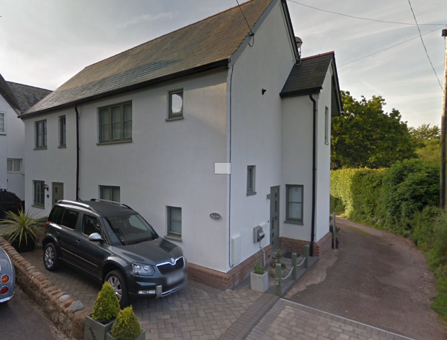 Exterior view of a white residential building with gray roof, parked black SUV, and driveway with small potted plants, next to a dirt path and greenery.
