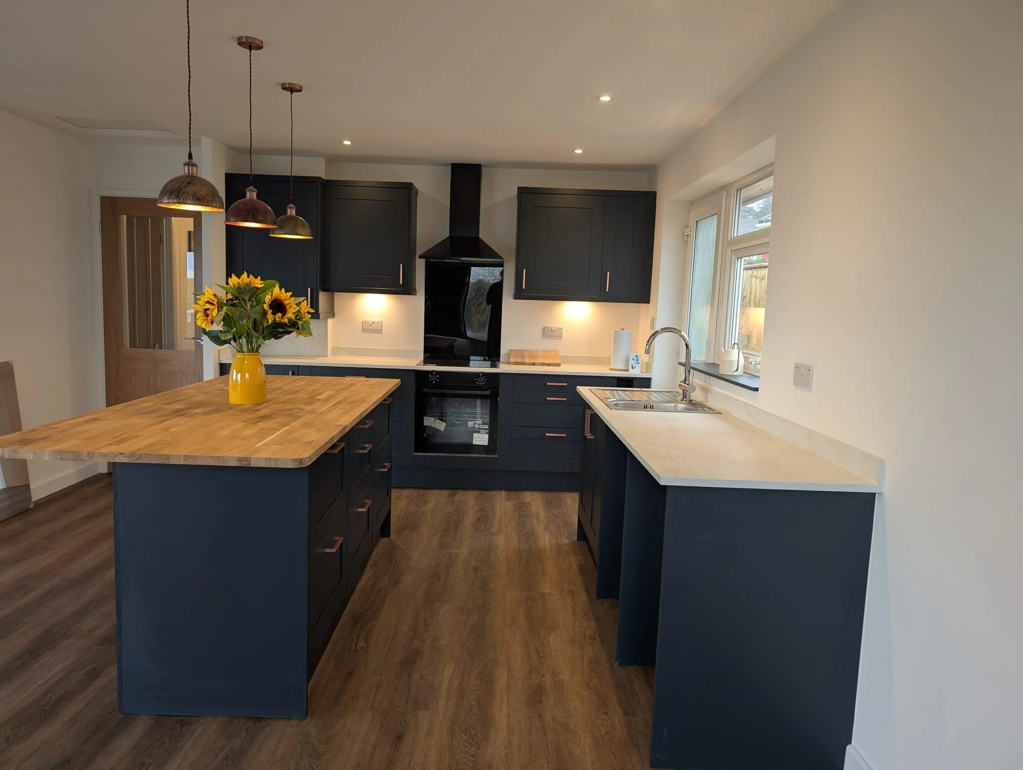 Modern kitchen with navy blue cabinets, wooden countertops, and a kitchen island with sunflower arrangement in a yellow vase. Includes black oven, range hood, window, and pendant lights.