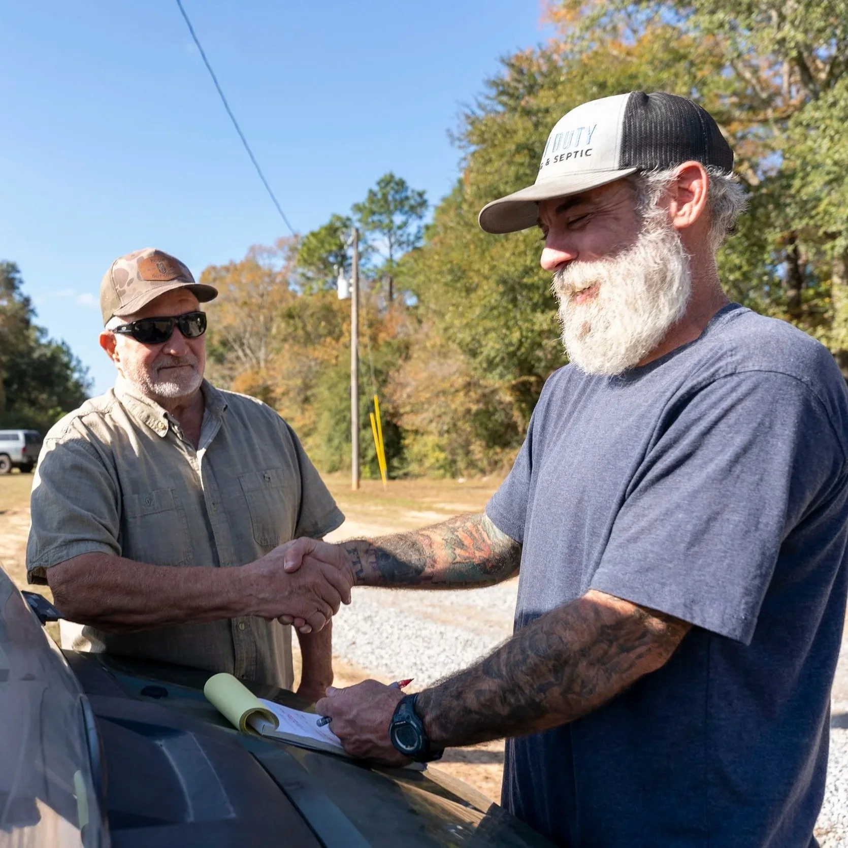 Two septic technicians shaking hands beside truck after residential tank service visit, discussing maintenance results outdoors on sunny rural property driveway.