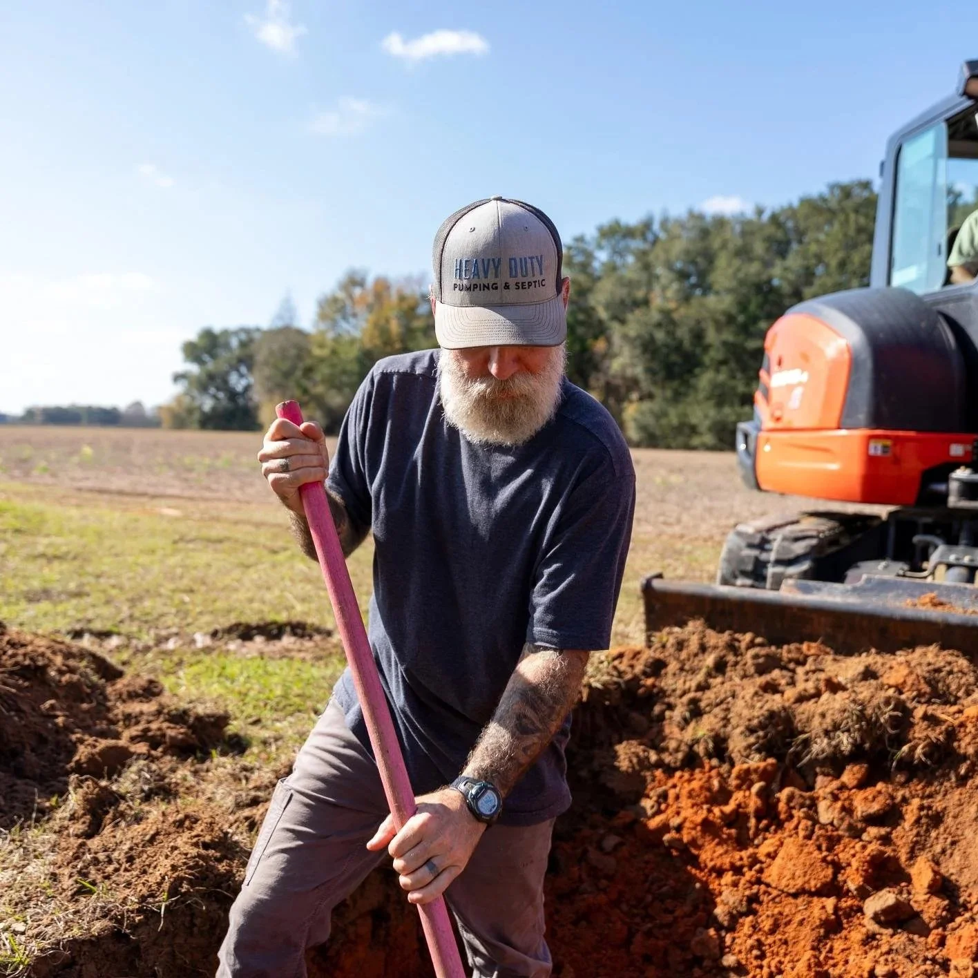 Septic technician digging beside residential drain field during system upgrade project after household expansion increased wastewater demand beyond original capacity.