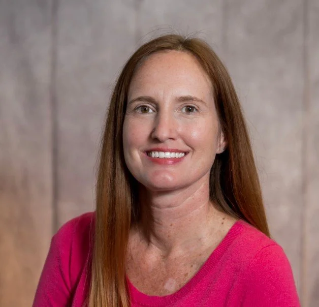 A woman with long red hair wearing a pink top, smiling, standing in front of a neutral background.