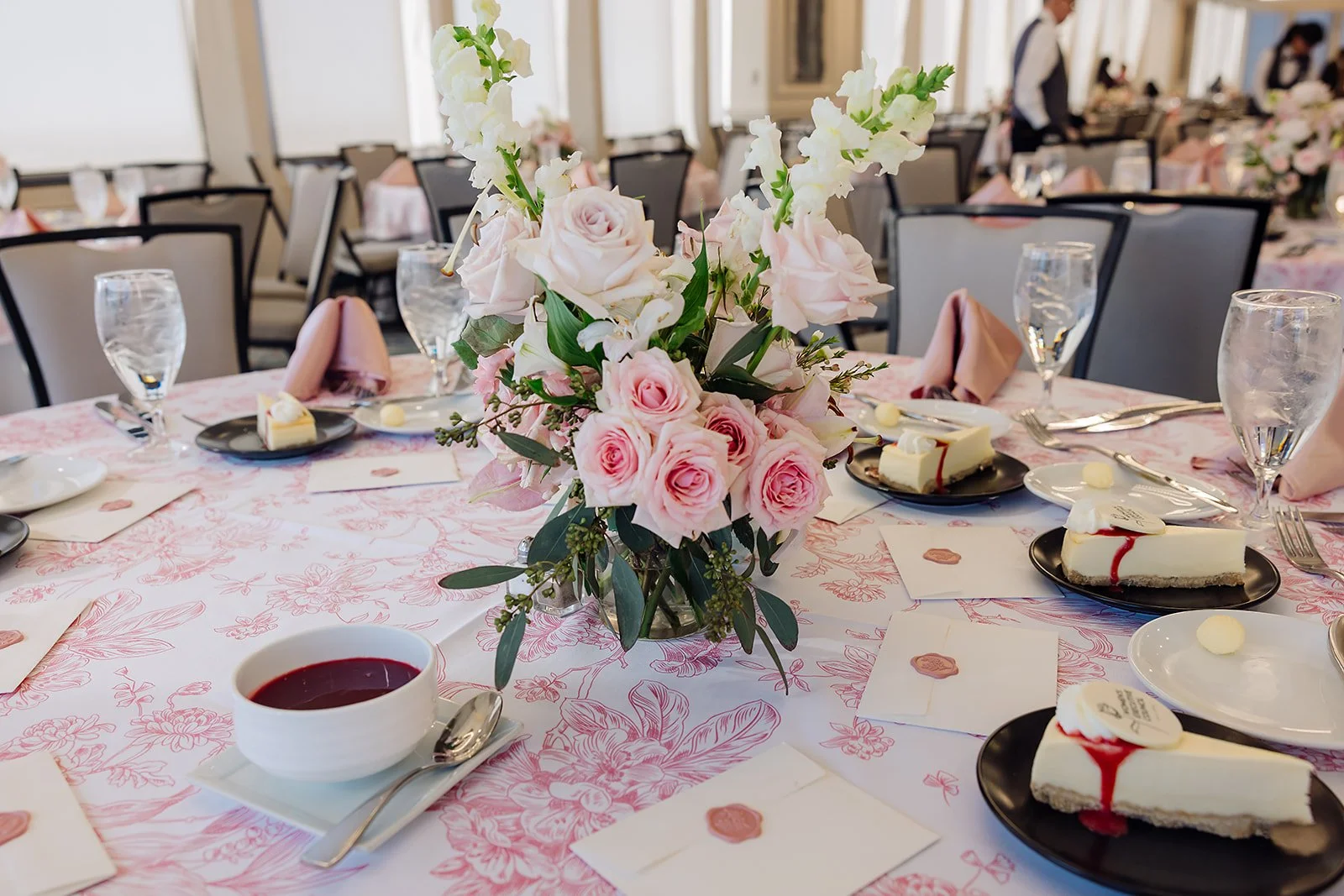 Elegant banquet table decorated with a pink floral centerpiece, pink napkins, wine glasses, and plates of cheesecake slices in a bright dining room.
