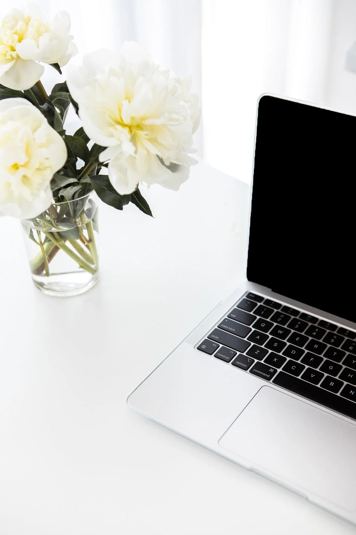 A laptop on a white desk next to a glass vase with white and yellow peony flowers.