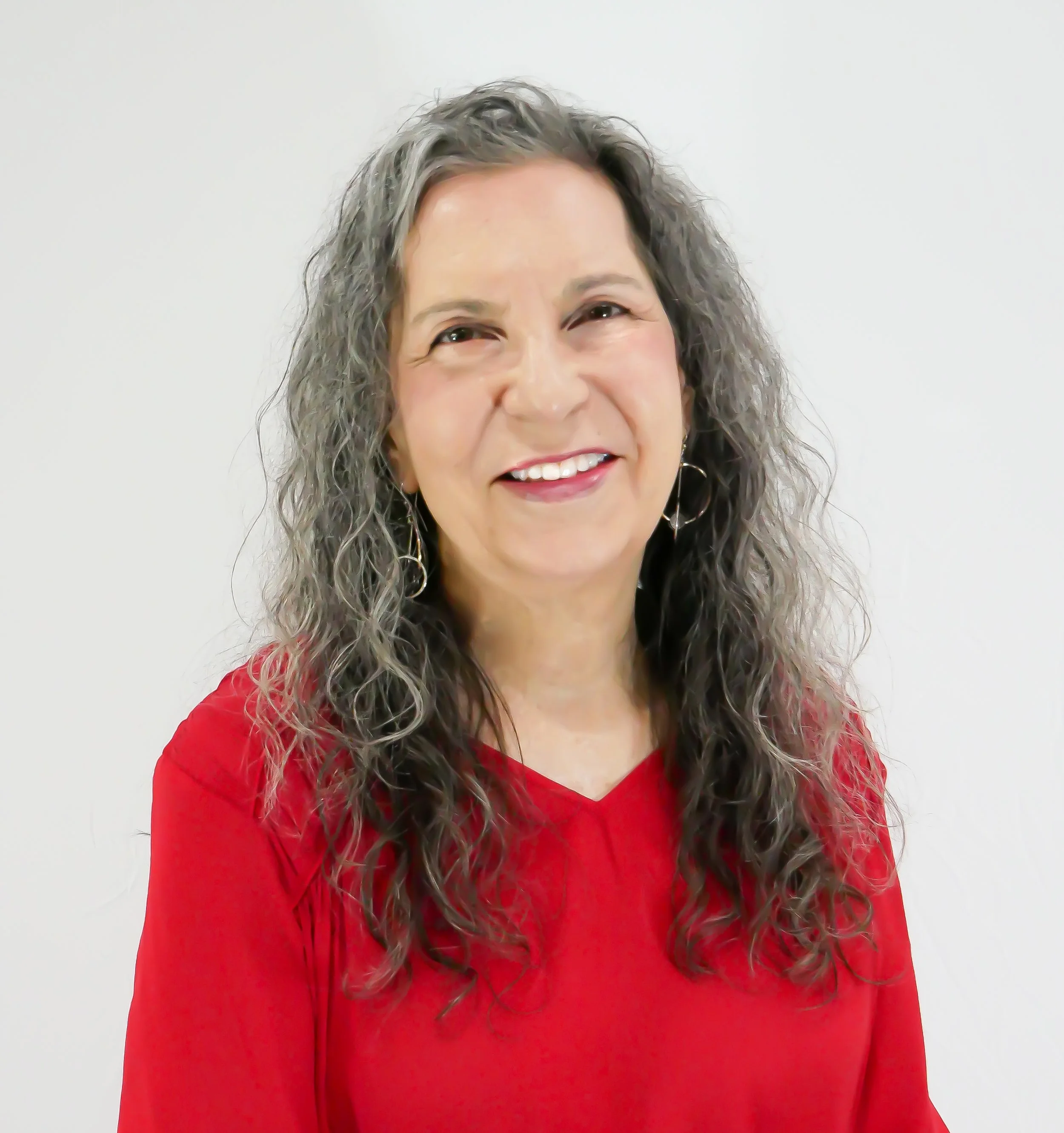 A woman with curly gray hair wearing a red top and hoop earrings, smiling against a plain white background.