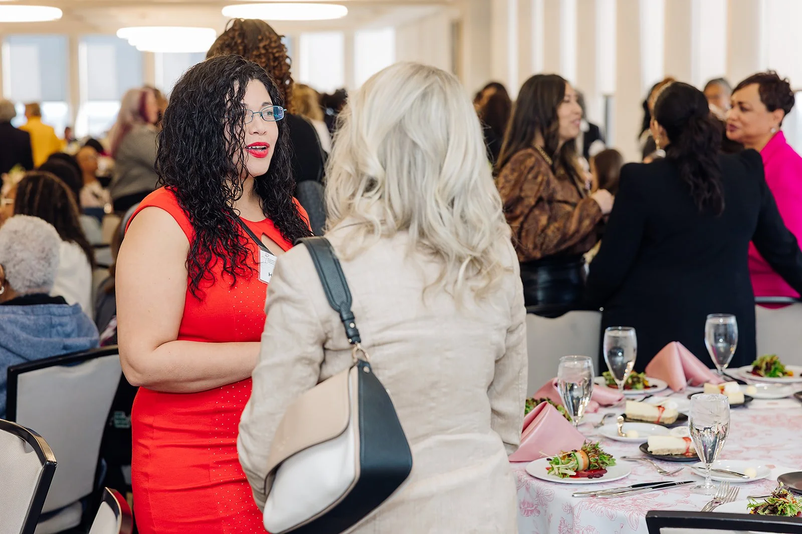 Women gathering and conversing at a formal event with tables set with plates, cutlery, glasses, and pink napkins.