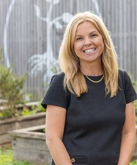 A woman with blonde hair wearing a black shirt, smiling outdoors in front of a concrete wall with plants.