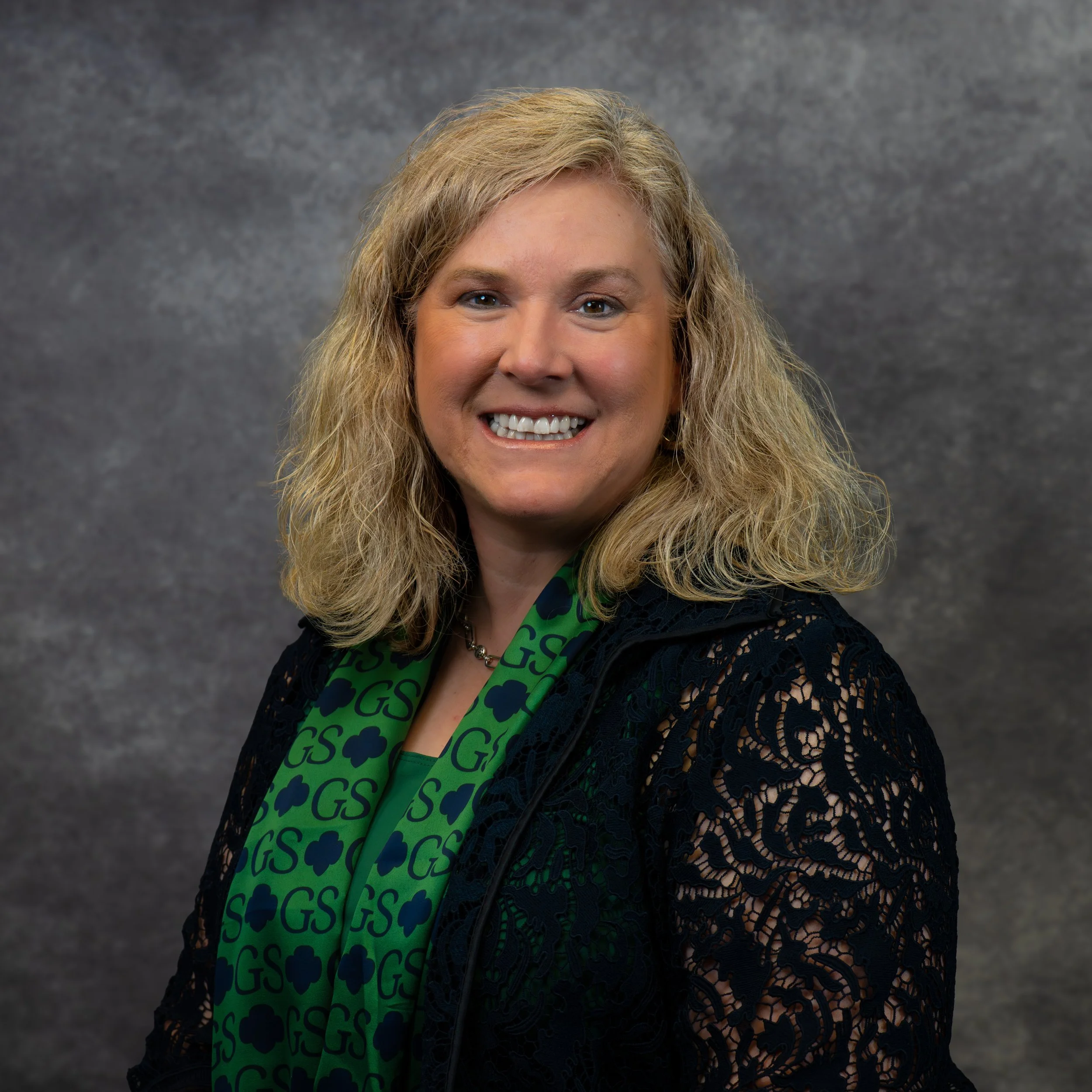 A woman with blonde, wavy hair, smiling, wearing a green scarf with shamrock and initials G S, and a dark lace top, against a gray textured background.
