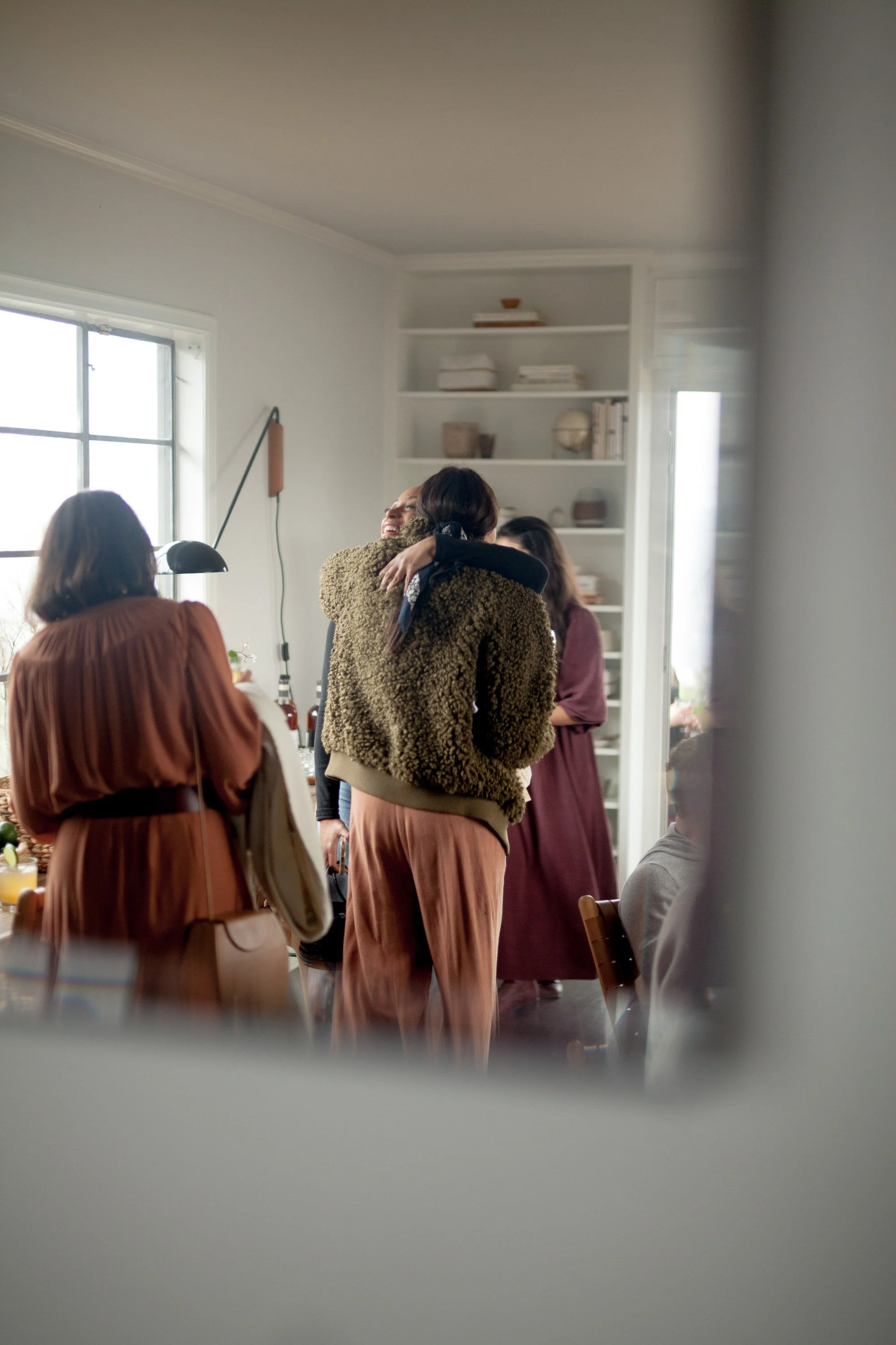 People hugging inside a well-lit room with white walls and shelves in the background.