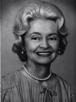 Black and white photo of a smiling woman with styled hair, pearl earrings, and a string of pearls, wearing a blouse with a bow tie detail.