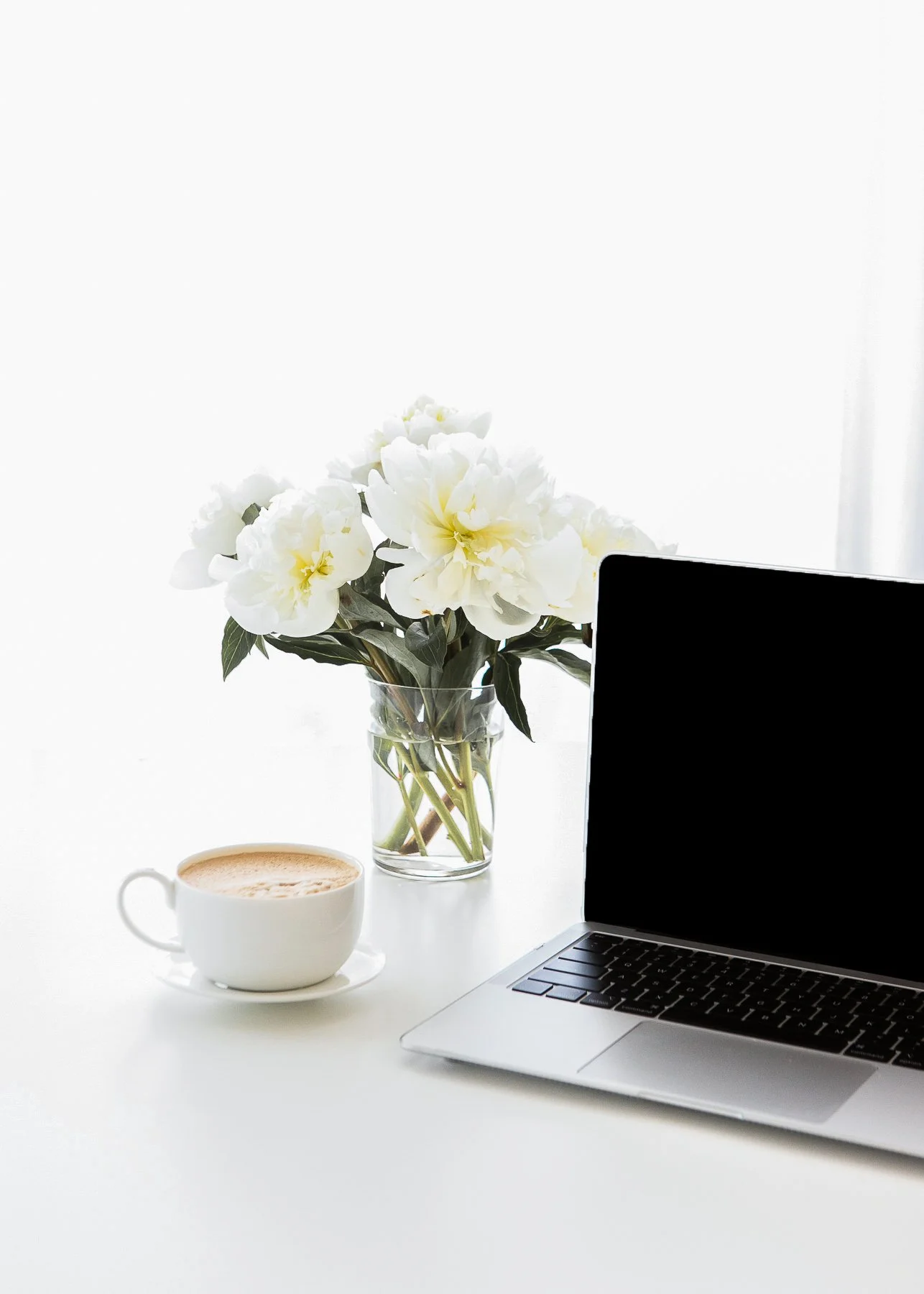 A laptop, a cup of coffee, and a vase of white flowers on a white desk