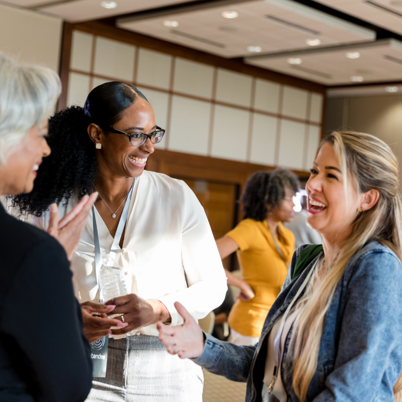 Three women engaged in a cheerful conversation at a networking event, with one woman shaking hands with another.