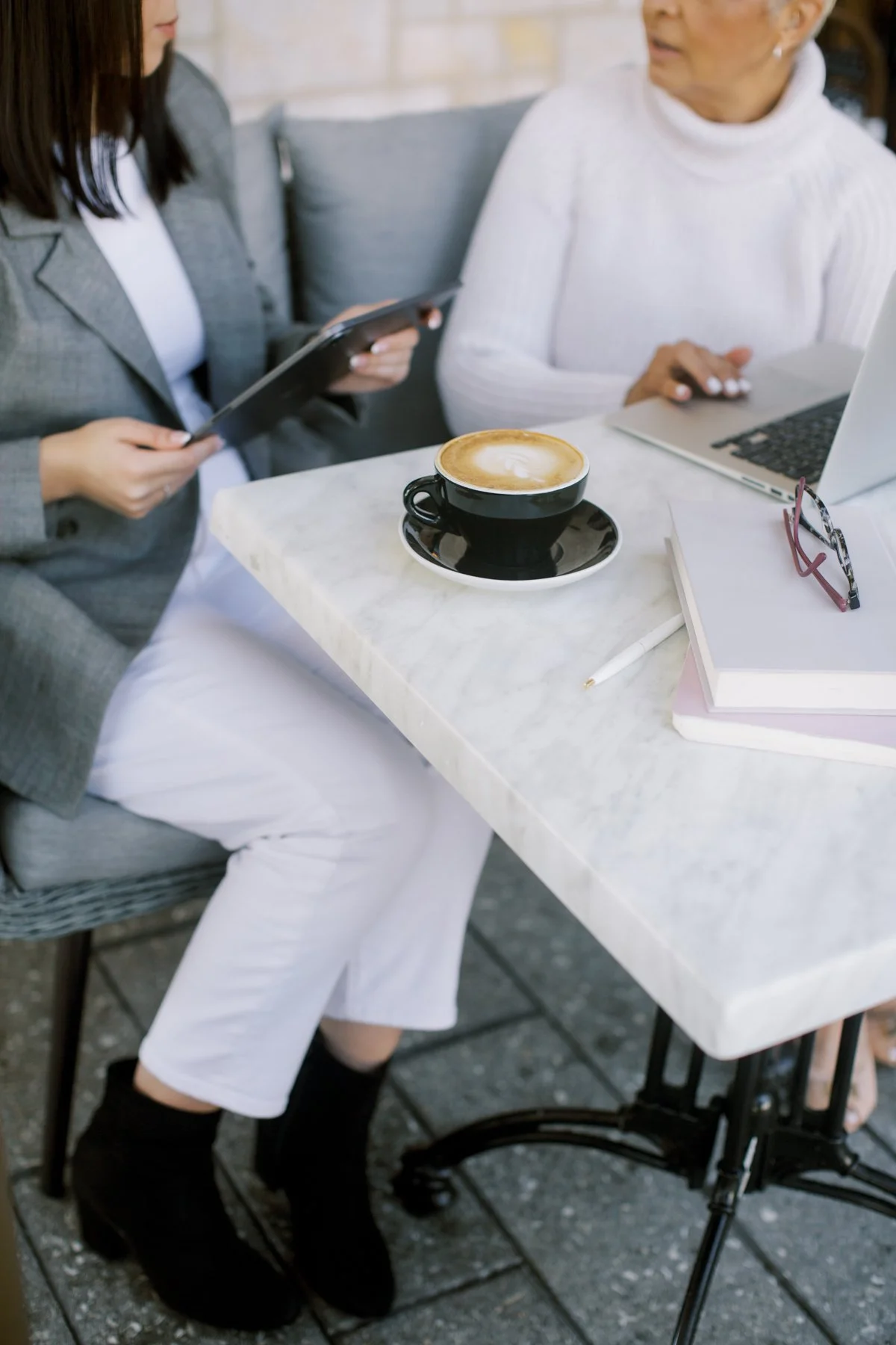 Two women sit at a marble table, one holding a tablet and the other typing on a laptop. There is a cup of coffee, a notebook, glasses, and a pen on the table. They are in a cozy indoor or outdoor setting.